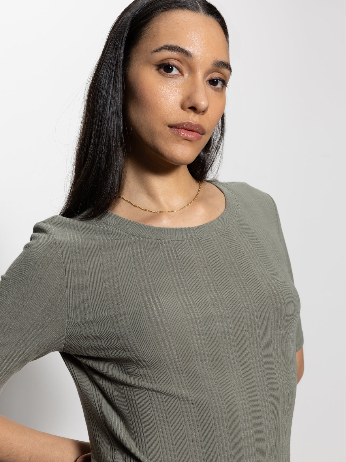 A long-haired individual with dark locks is wearing the Sanctuary Clothing half sleeve femme crew tee in dark spruce and a thin gold necklace, gazing at the camera against a plain white background.