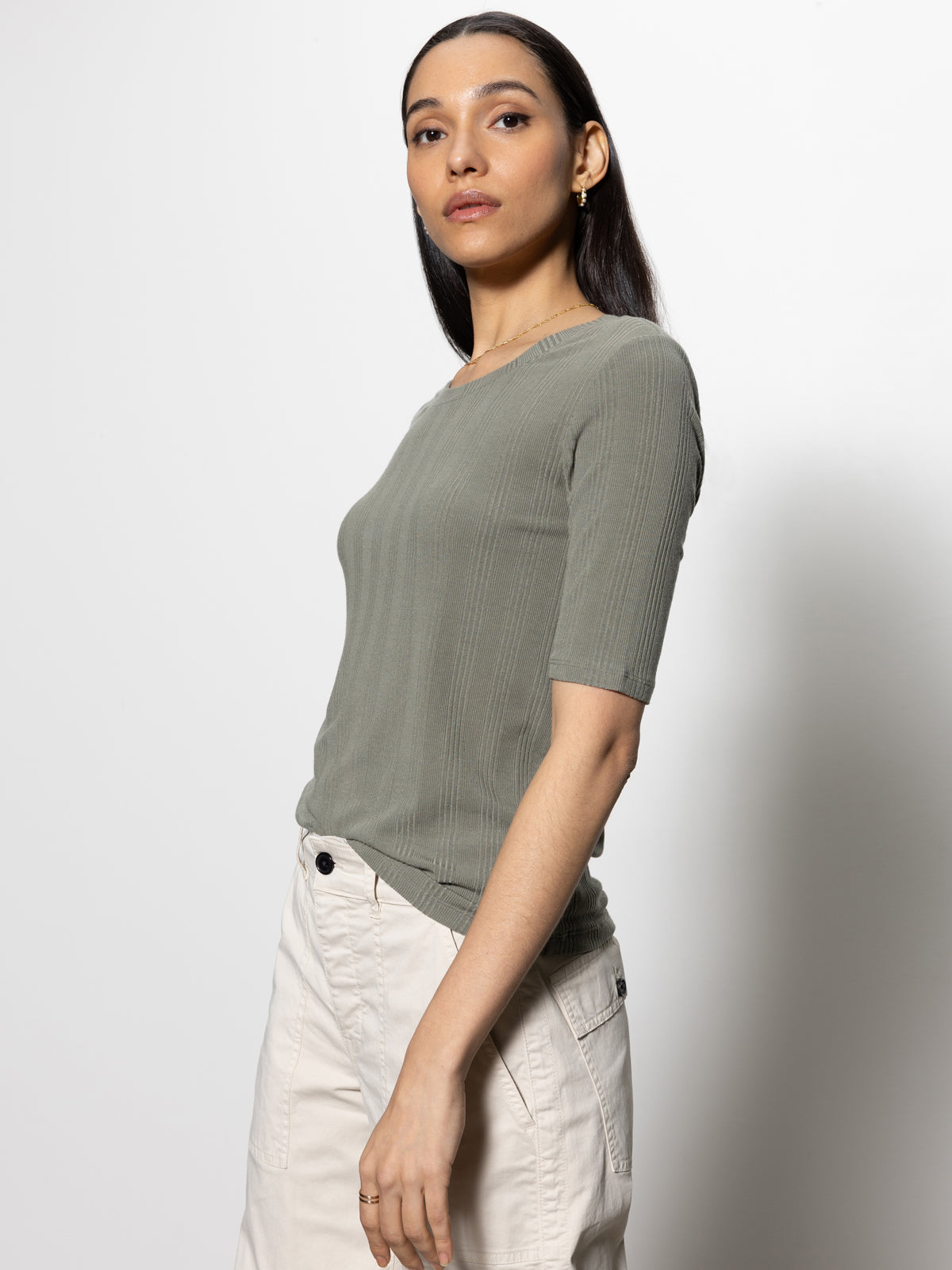 A person with long dark hair wears a Sanctuary Clothing half sleeve femme crew tee in dark spruce and off-white pants. They stand against a plain white background, facing slightly to the side while looking at the camera.