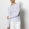 A dark-haired person is wearing a Sanctuary Clothing long sleeve boatneck t-shirt in sky blue and white pants. They stand against a plain white background, looking forward calmly, with their right arm crossed over their body.