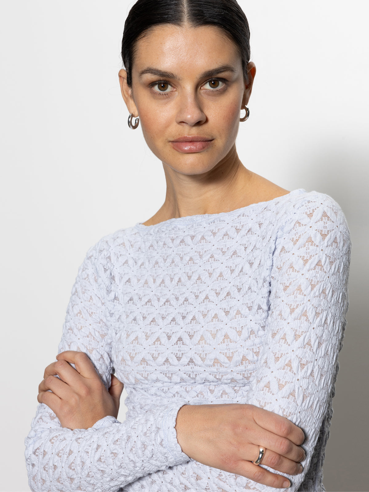 A person with dark hair pulled back, in hoop earrings and a Sanctuary Clothing long sleeve boatneck T-shirt in sky blue, stands with arms crossed against a plain backdrop. Their expression is neutral as they look directly at the camera.