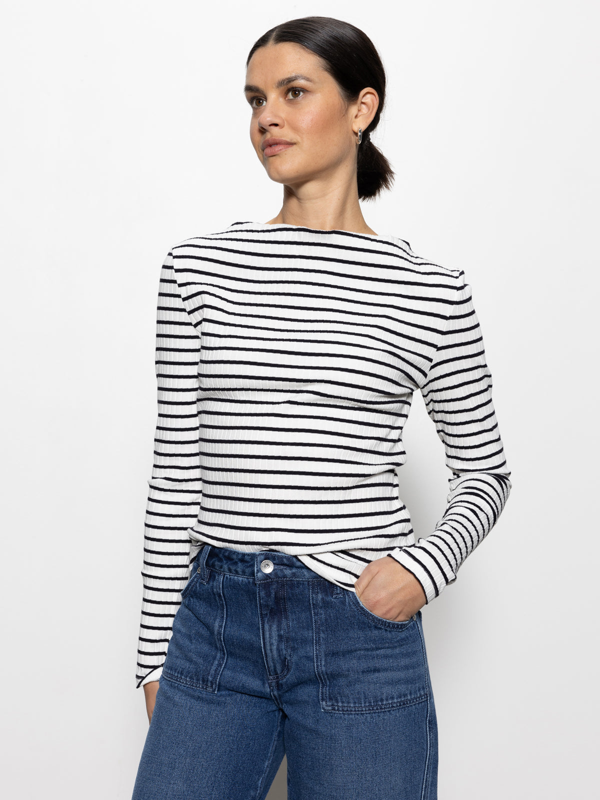 A person with dark hair pulled back is wearing a Sanctuary Clothing long sleeve boatneck t-shirt in chalk black stripe and blue jeans. They stand against a plain white background, one hand in a pocket, looking to the side.