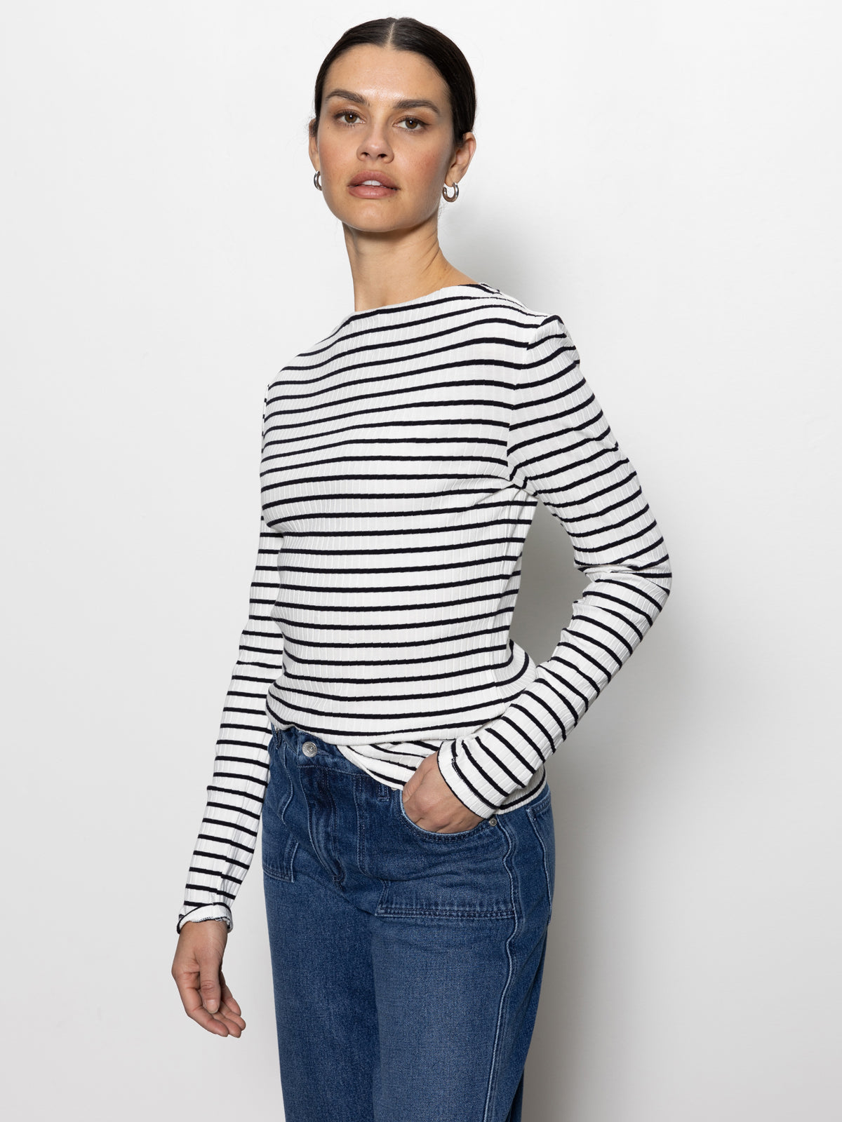 A person in a Sanctuary Clothing long sleeve boatneck t shirt in chalk black stripe and blue jeans poses against a white background. Their right hand is in their pocket, and with short, dark hair, they gaze confidently at the camera.