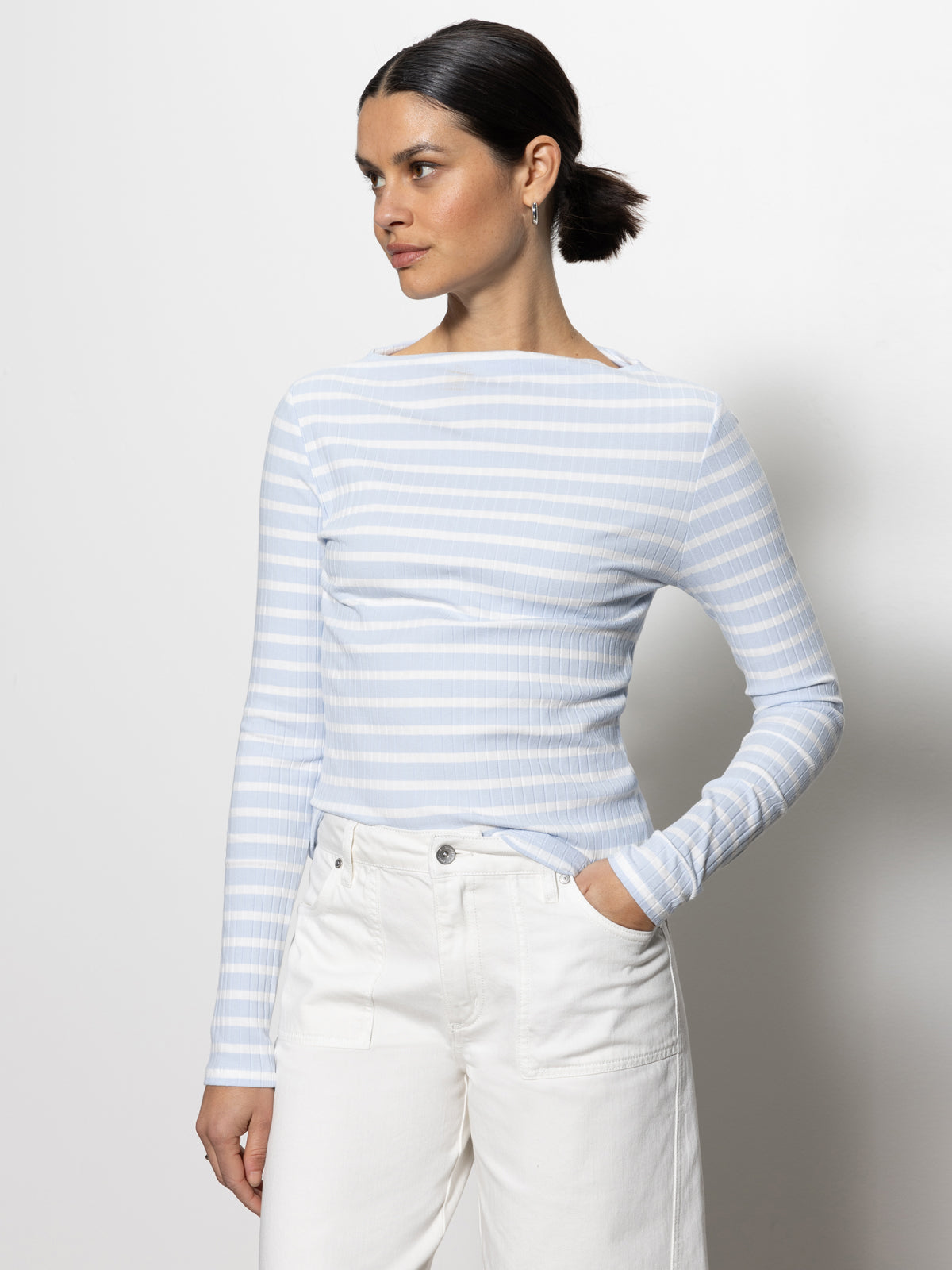 A person with dark hair in a low bun wears Sanctuary Clothing's long sleeve boatneck T-shirt in sky blue and white stripes, paired with white pants. They stand against a plain white background, looking to the side with one hand in a pocket.