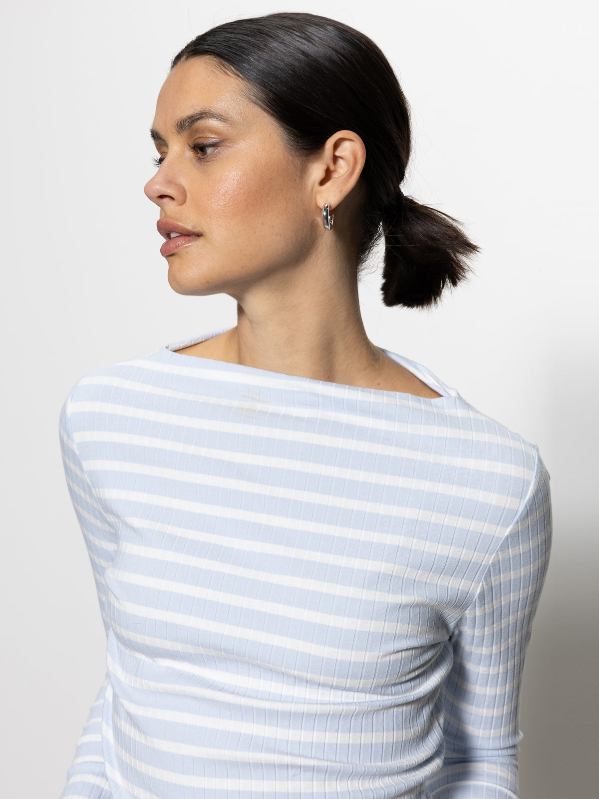 A person with dark hair tied back is wearing a long sleeve boatneck t-shirt in sky blue and white stripe by Sanctuary Clothing, along with small hoop earrings. They are looking to the side against a plain white background.