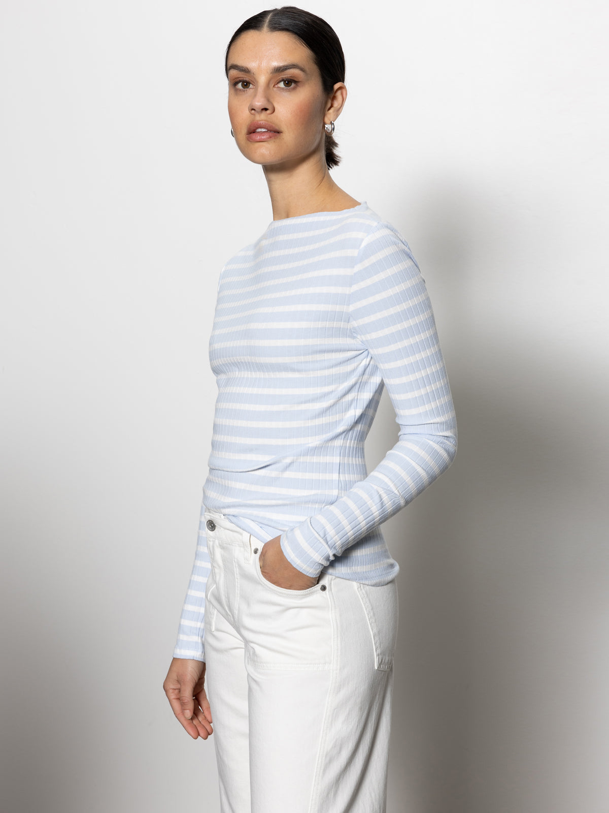 A woman in a Sanctuary Clothing long sleeve boatneck t-shirt, featuring sky blue and white stripes, paired with white pants against a plain background. Her hand rests in her pocket as she gazes slightly to the side.