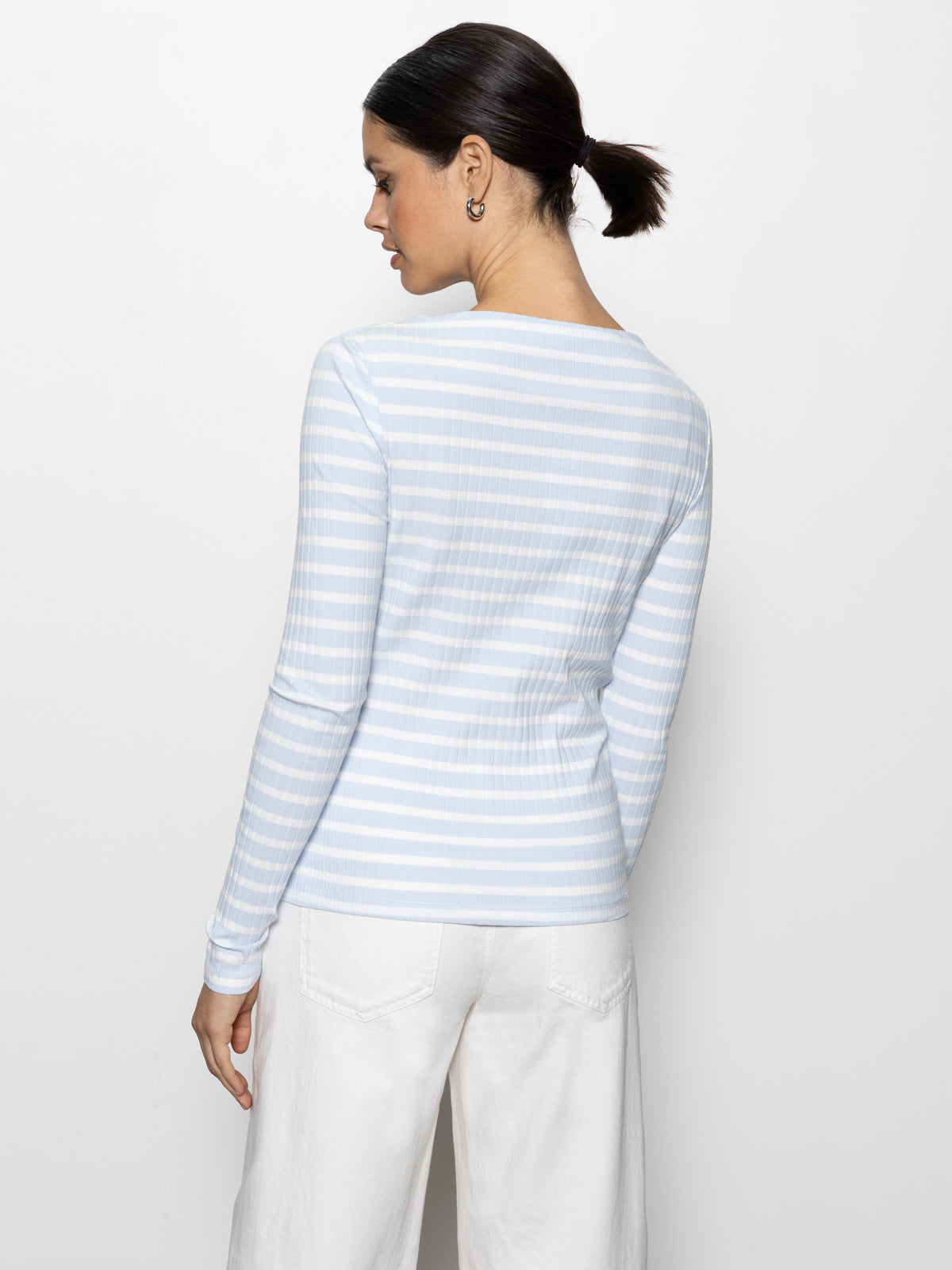 A woman with dark hair in a ponytail stands against a plain background, wearing Sanctuary Clothing's long-sleeve boatneck t-shirt in sky blue and white stripes, paired with white pants as she gazes sideways.