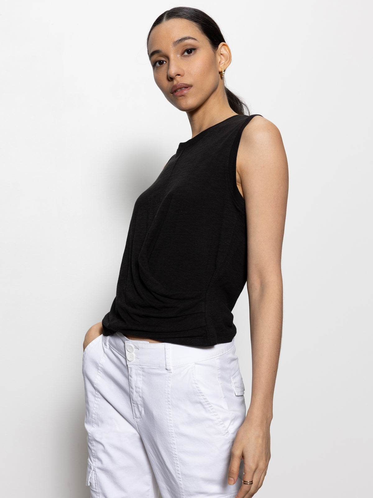 A woman stands against a white background, gazing at the camera with a relaxed expression. Her long hair is tied back as she wears the sleeveless draped tank in black by Sanctuary Clothing paired with white pants, her left hand resting in her pocket.