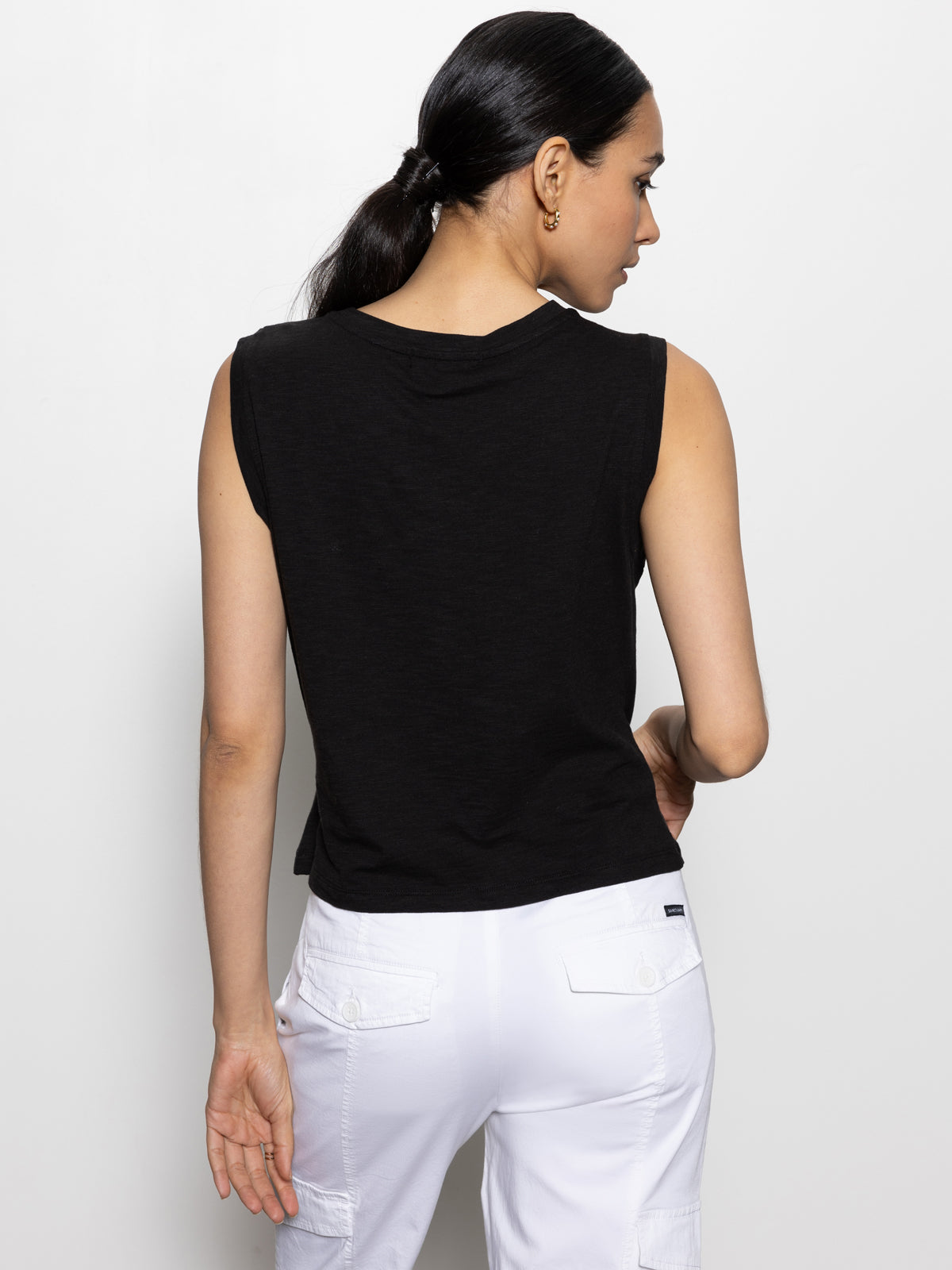 A person with long dark hair in a ponytail, wearing Sanctuary Clothing’s draped tank in black and white pants with pockets, is facing away from the camera against a plain white background.