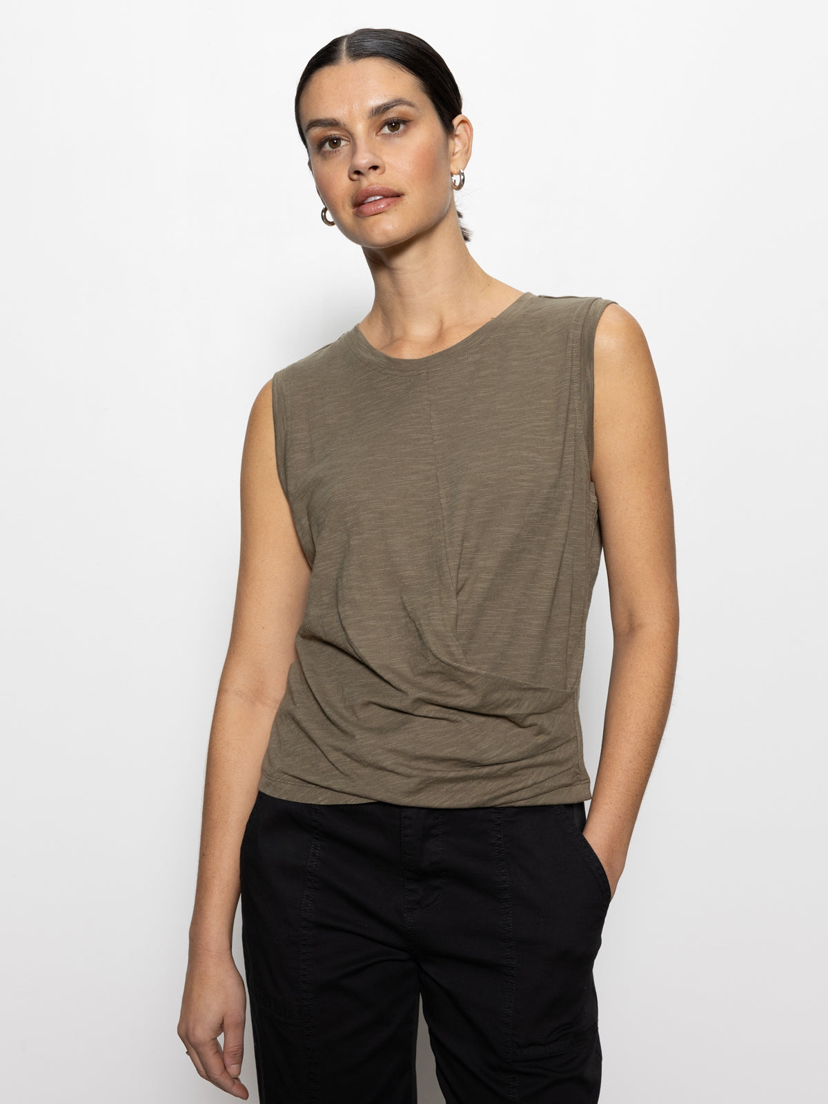 A person in a Sanctuary Clothing draped tank in burnt olive and black pants stands against a plain white background. Their dark hair is pulled back, they wear hoop earrings, and have a neutral expression.