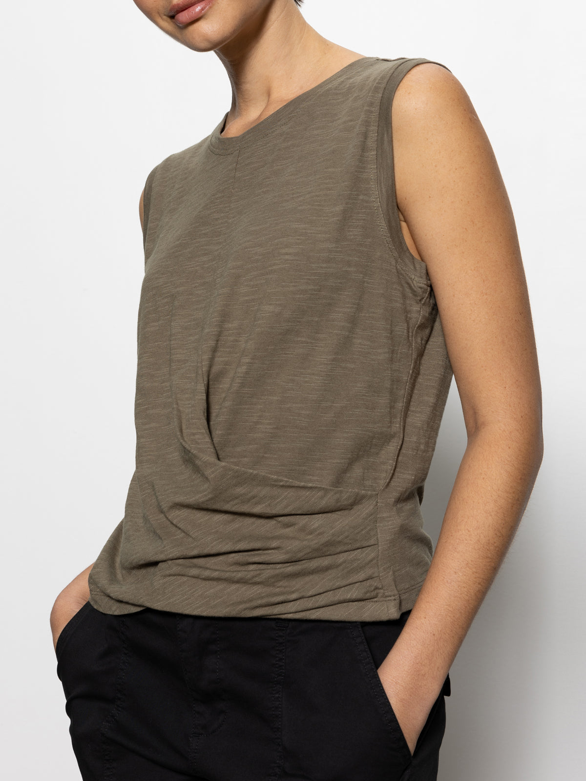 A person in a Sanctuary Clothing draped tank burnt olive and black pants stands against a white background, with their head partially out of frame and one hand tucked into a pocket.