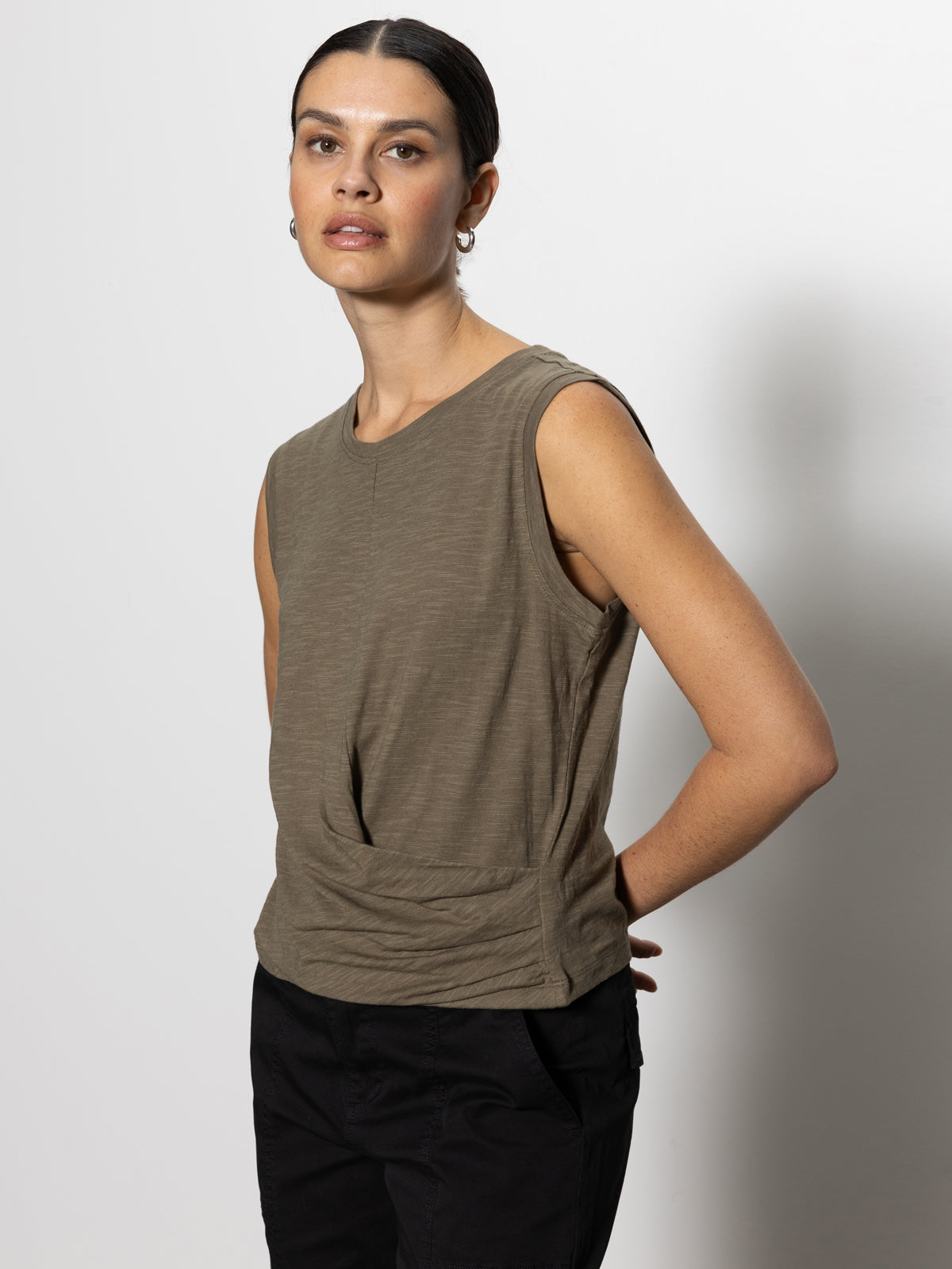 A person with dark hair tied back is wearing the Sanctuary Clothing draped tank in burnt olive with black pants. They pose against a white background, one hand on their hip, with a neutral expression.