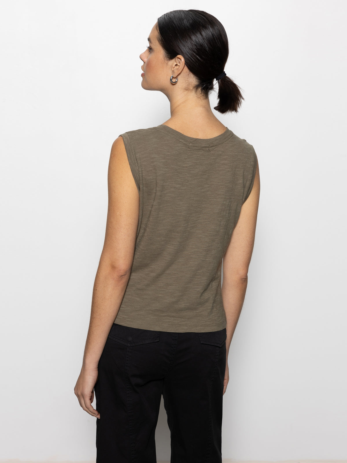 A person with dark hair in a ponytail showcases the back of their Sanctuary Clothing draped tank in burnt olive, paired with black pants, against a plain white background.