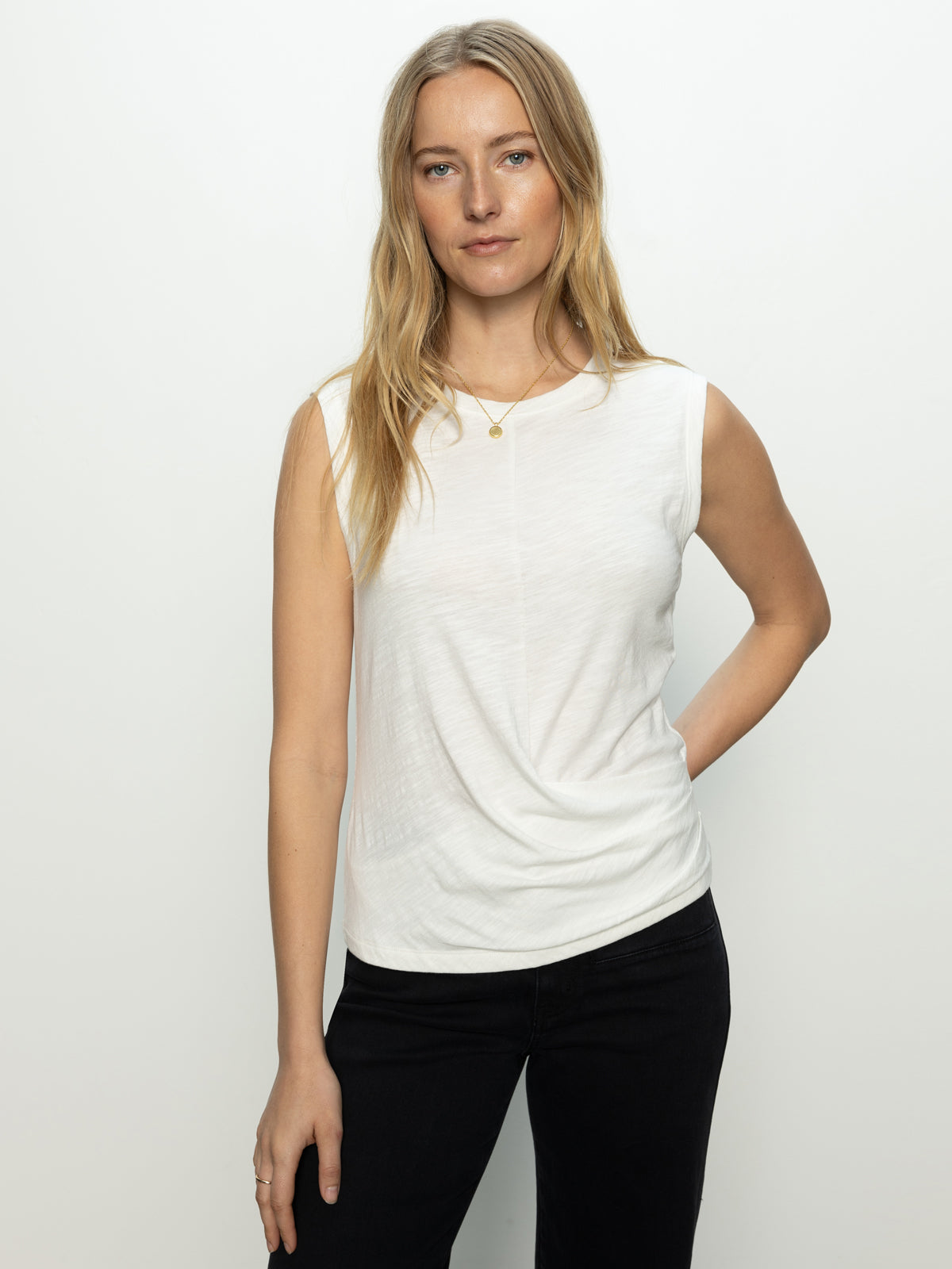 A woman with long blond hair wears Sanctuary Clothing's draped tank in white and black pants, standing against a plain background. With one hand on her hip and a necklace, she looks directly at the camera with a relaxed expression.