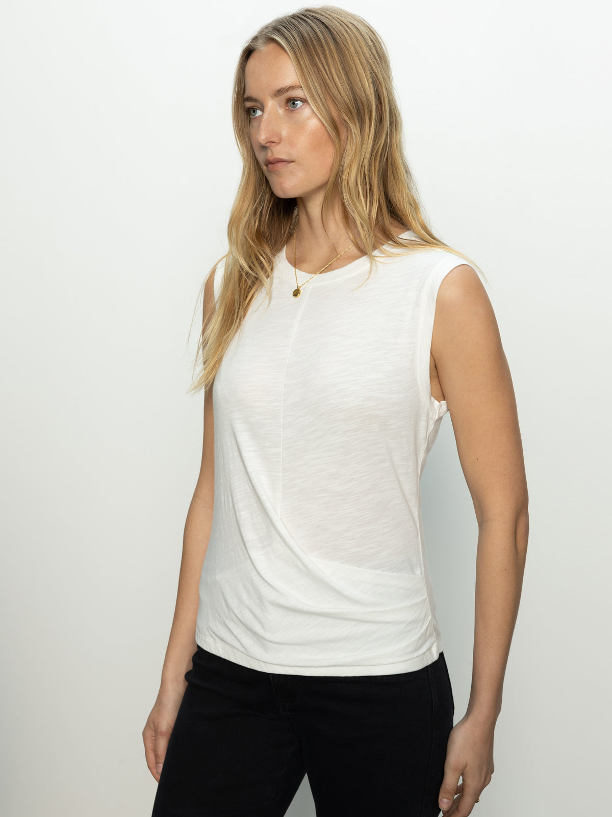 A woman with long blonde hair wears the draped tank white from Sanctuary Clothing, paired with black pants. She stands against a plain white background, looking off to the side.