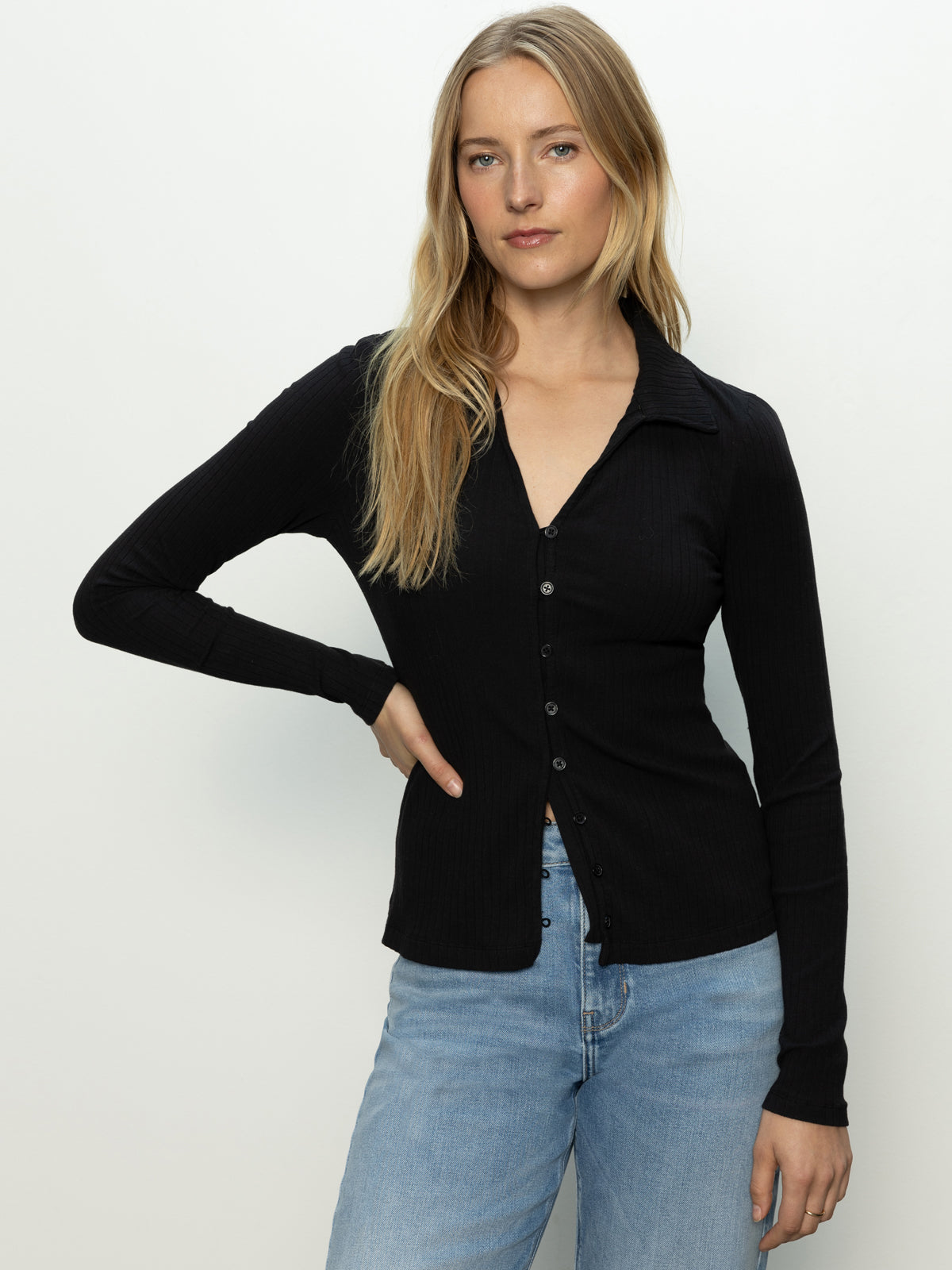 A person with long blonde hair wears a Sanctuary Clothing daydreamer knit shirt in black and light blue jeans, standing against a plain light background. They pose relaxedly with one hand on their hip.