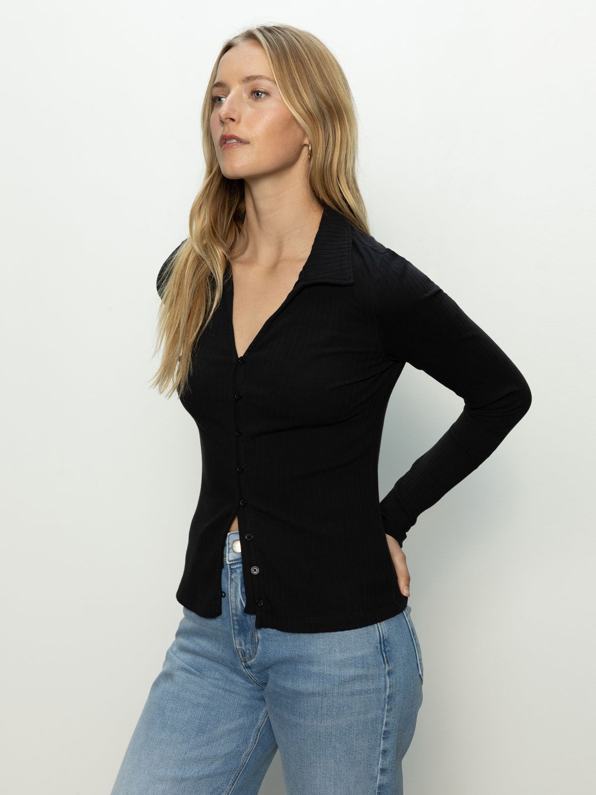 A woman with long blonde hair wears the Sanctuary Clothing daydreamer knit shirt in black and light blue jeans. She stands against a plain, light-colored background, gazing slightly to her left with a neutral expression.