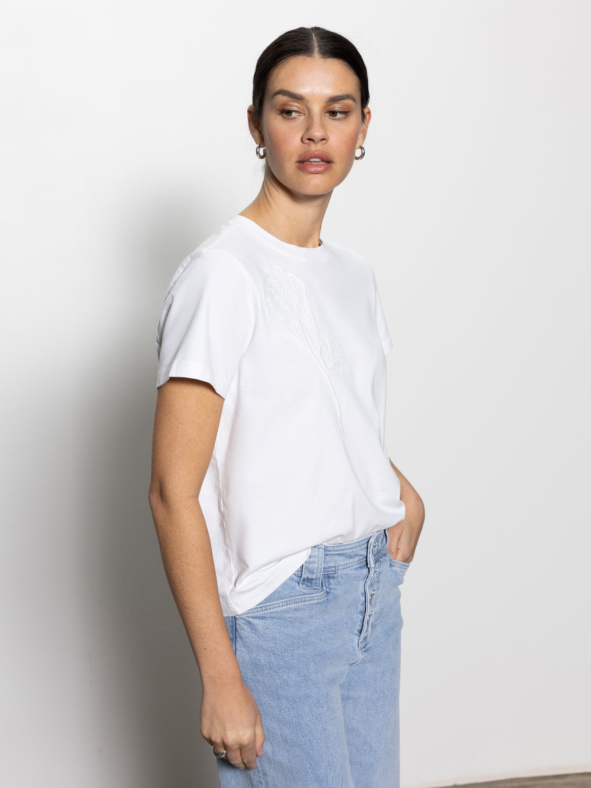 A person with dark hair is slightly turned to the side, wearing the rose perfect tee white by Sanctuary Clothing and light blue jeans. They have one hand in their pocket and a neutral expression, set against a plain white background.