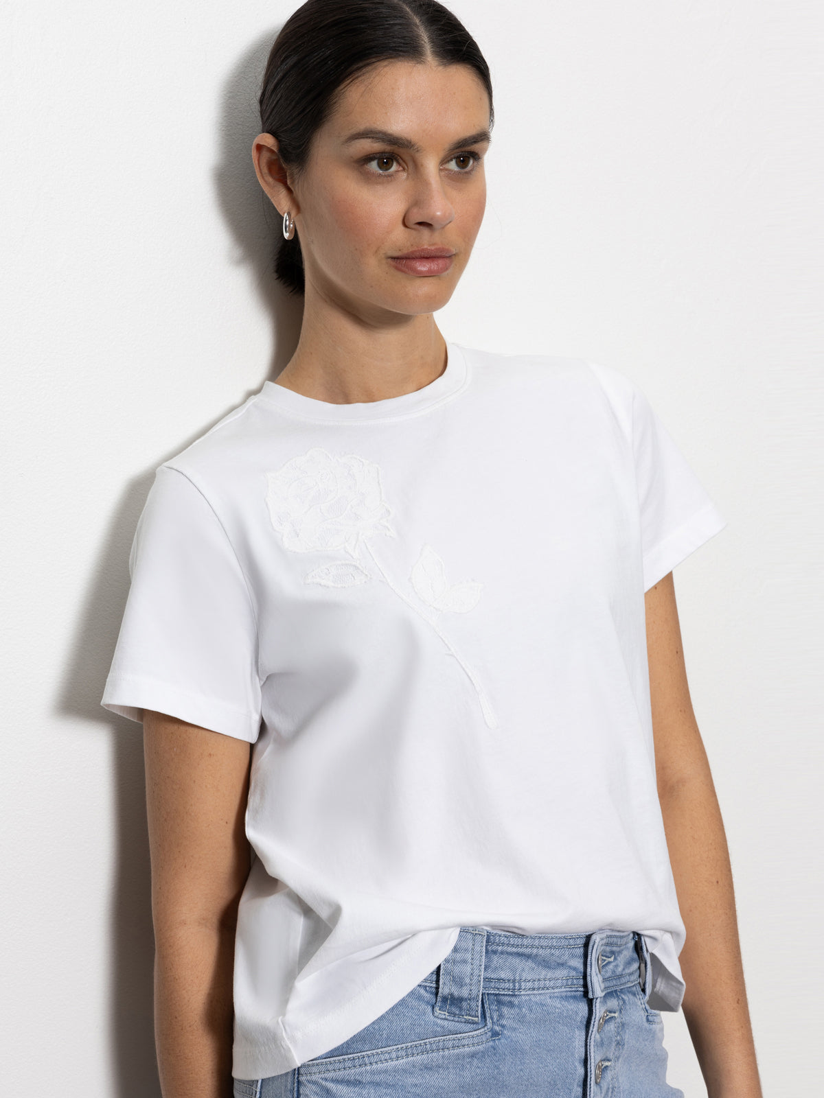 A woman stands against a white wall, wearing the "Rose Perfect Tee White" by Sanctuary Clothing. Her dark hair is pulled back, and she is paired with light blue jeans.