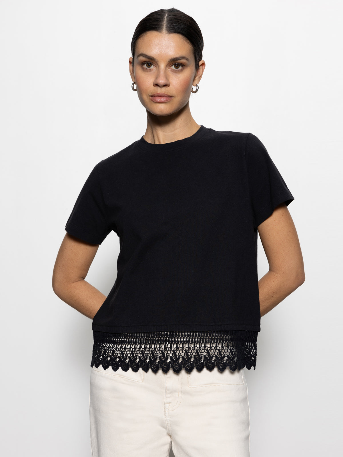 A person stands against a plain white background wearing Sanctuary Clothing's lace remix tee in black with lace detailing along the hem and white pants. They have short, dark hair and are wearing small hoop earrings.