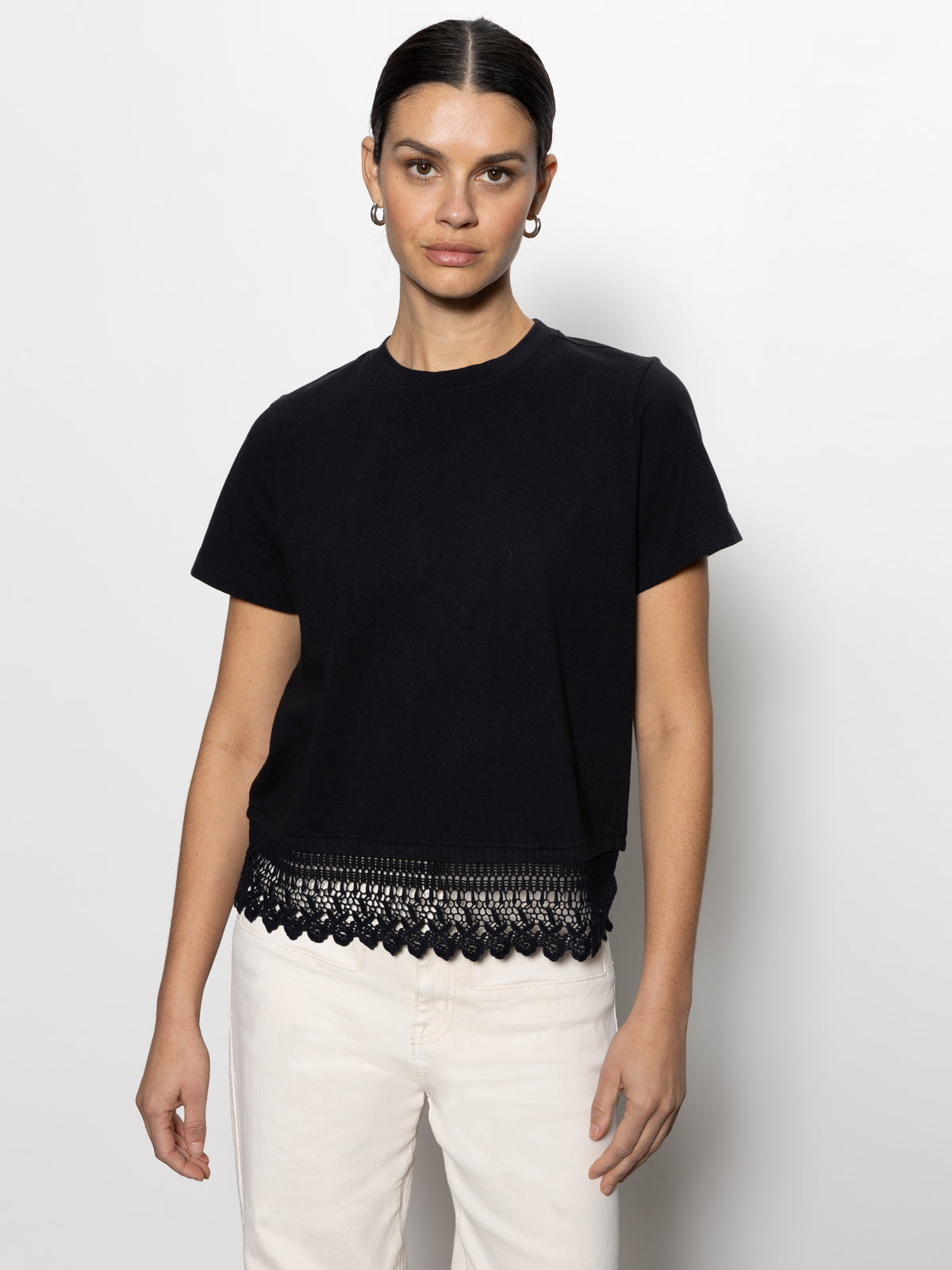 A person faces the camera wearing a "lace remix tee black" by Sanctuary Clothing with lace detailing at the hem, paired with white pants. The backdrop is plain and light-colored.