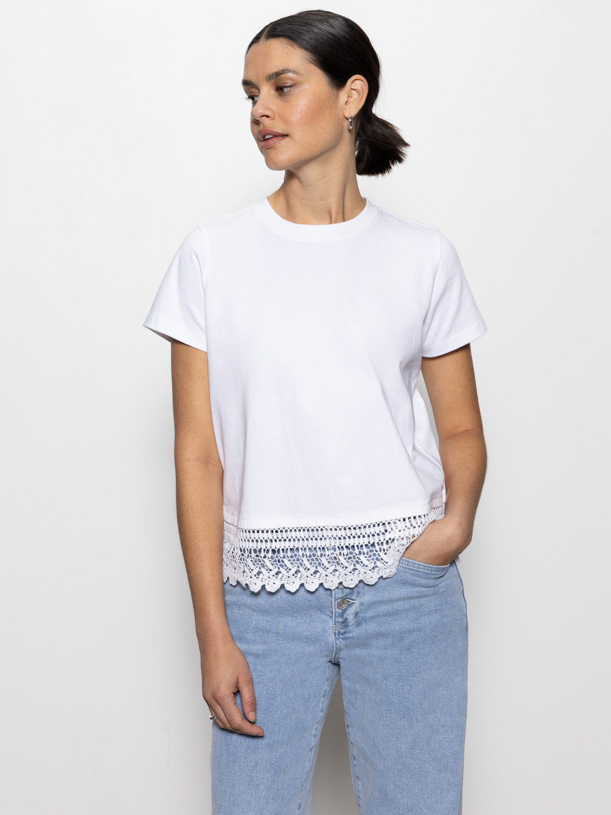 A woman with dark hair in a ponytail wears Sanctuary Clothing's "Lace Remix Tee" in white and light blue jeans. She stands against a plain white background, hand in pocket, looking to the side.