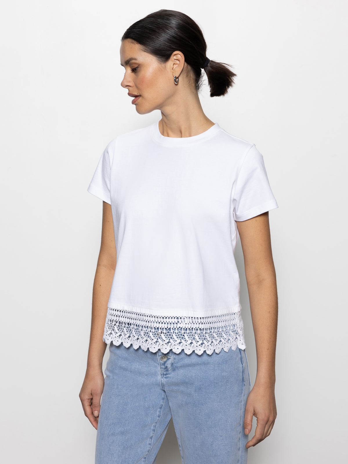 A woman with her hair in a low bun wears the lace remix tee by Sanctuary Clothing and light blue jeans. She looks to her left against a plain white background.