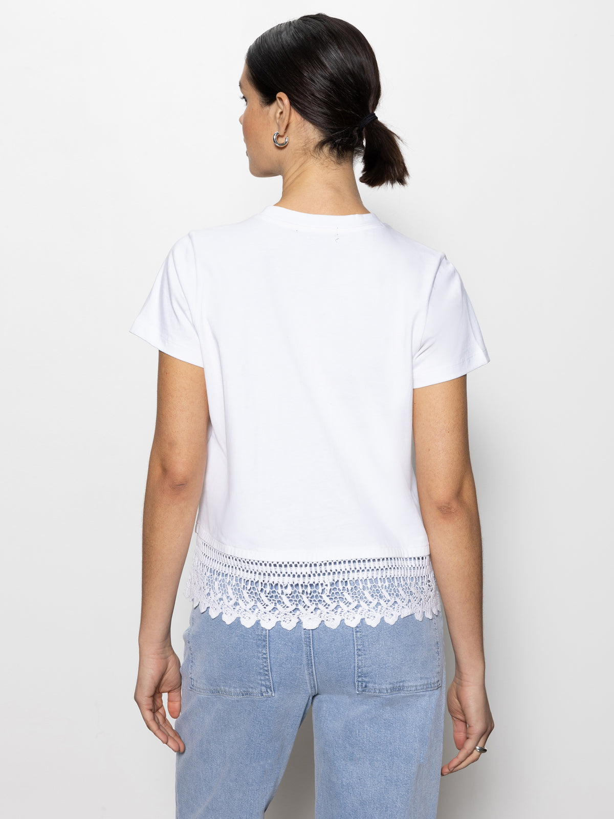 A person with dark hair in a ponytail wears a Sanctuary Clothing lace remix tee in white paired with light blue jeans, standing against a white backdrop.