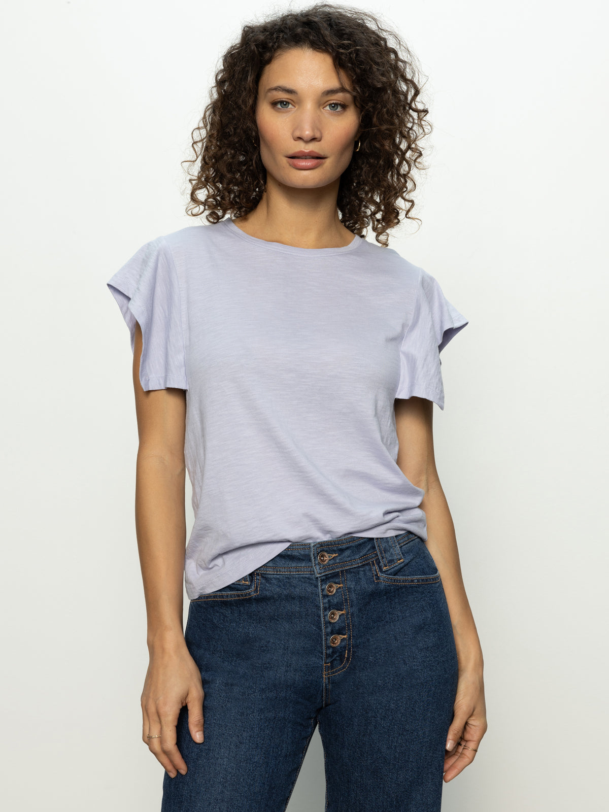 A person with curly hair stands against a plain background, wearing Sanctuary Clothing's relaxed muscle tee in lavender mist and blue jeans with a button-up fly, looking forward with a neutral expression.