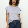 A person with curly hair stands against a plain background, wearing Sanctuary Clothing's relaxed muscle tee in lavender mist and blue jeans with a button-up fly, looking forward with a neutral expression.