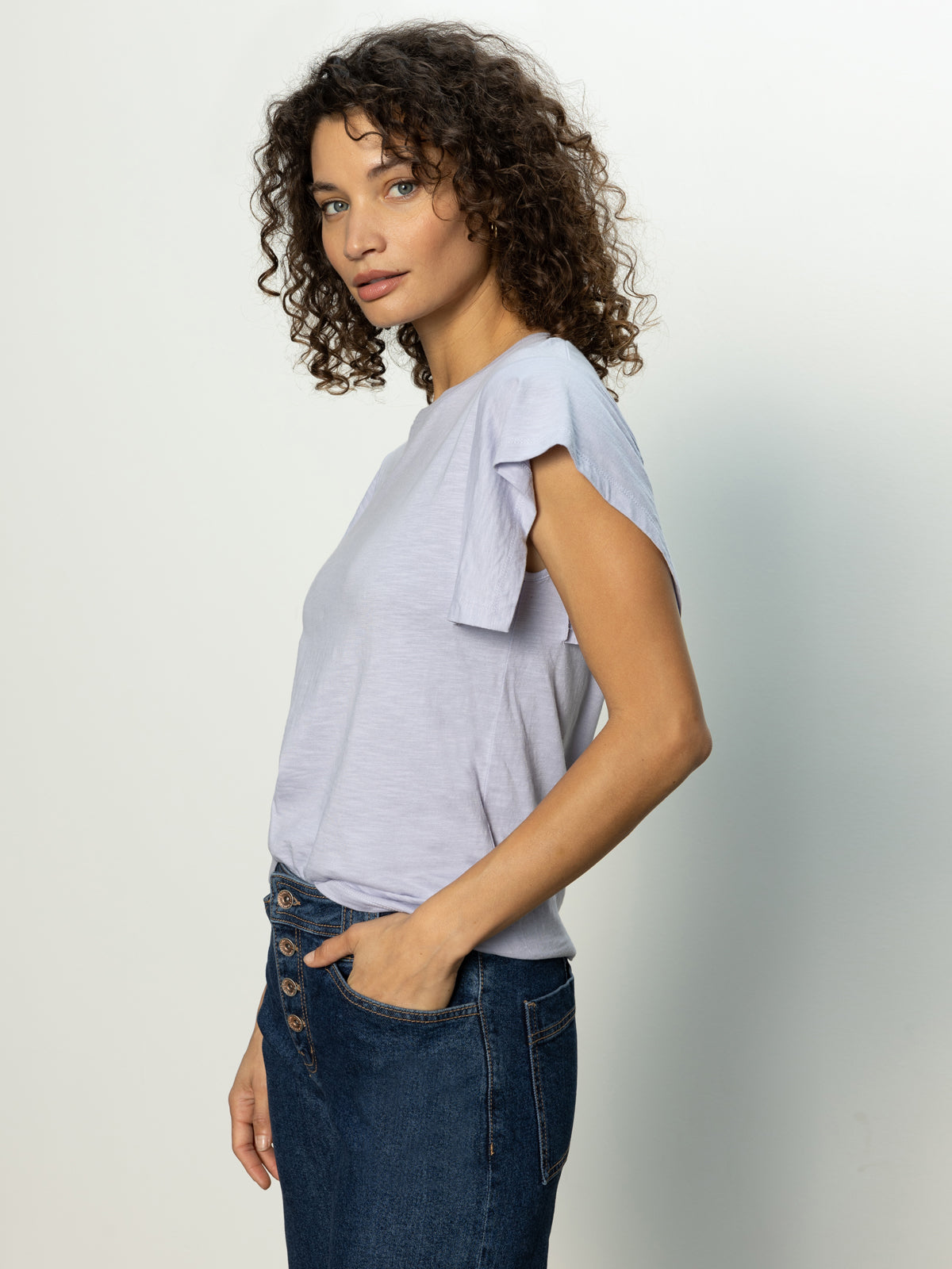 A person with curly hair wearing Sanctuary Clothing's Relaxed Muscle Tee in Lavender Mist and blue jeans stands sideways, their right hand in the pocket, against a plain light-colored background.