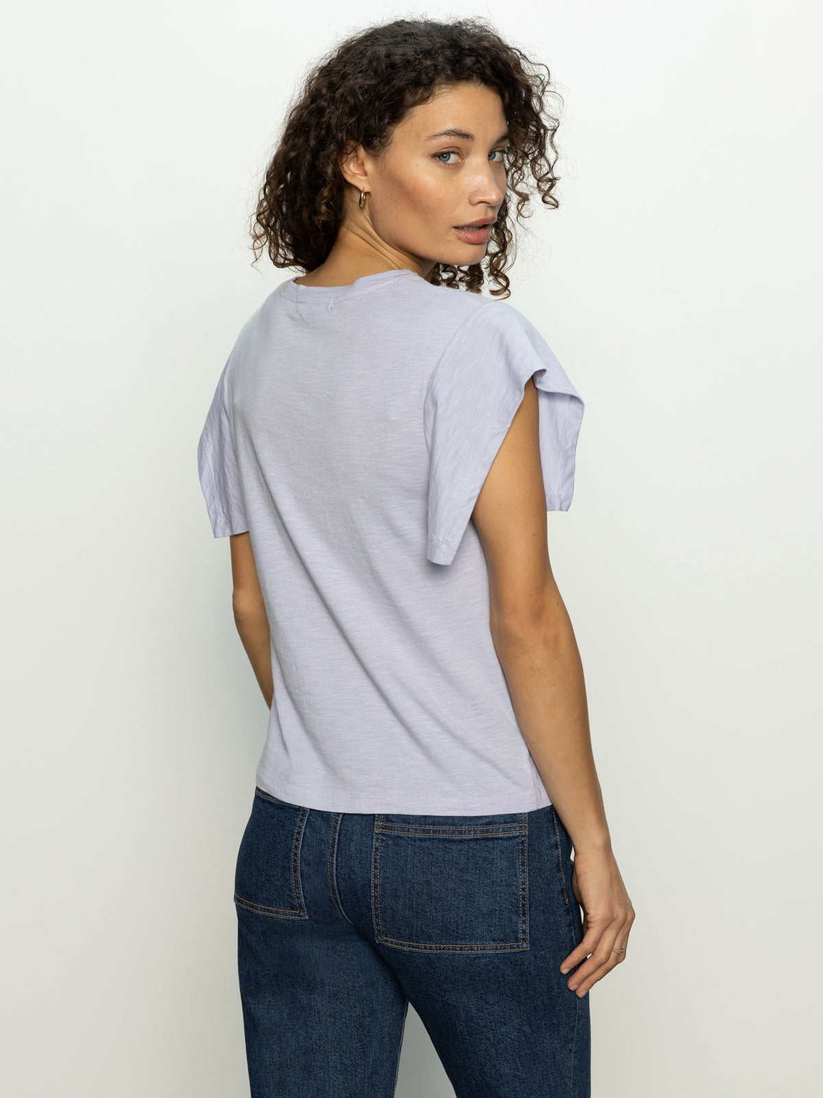 A woman with curly hair is standing and looking over her shoulder, wearing a Sanctuary Clothing relaxed muscle tee in lavender mist and dark blue jeans against a plain light-colored background.