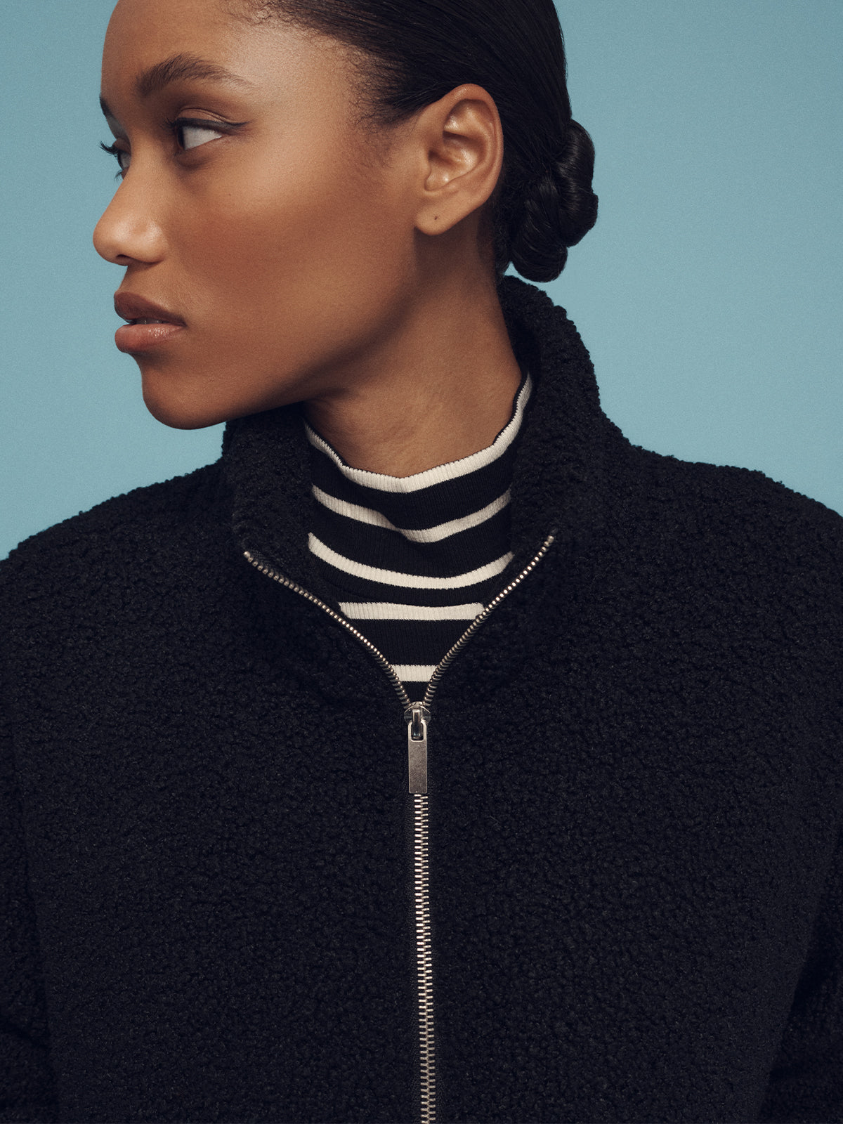 A person with their hair pulled back in a bun looks to the side against a blue backdrop, wearing the Sanctuary Clothing black alpine sherpa half zip popover over a black and white striped turtleneck.