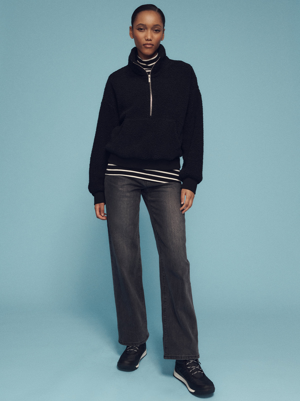 A person stands against a blue background wearing an alpine sherpa half zip popover in black by Sanctuary Clothing, along with a striped shirt, dark jeans, and black sneakers. They have short hair and a confident expression, with their hands by their sides.