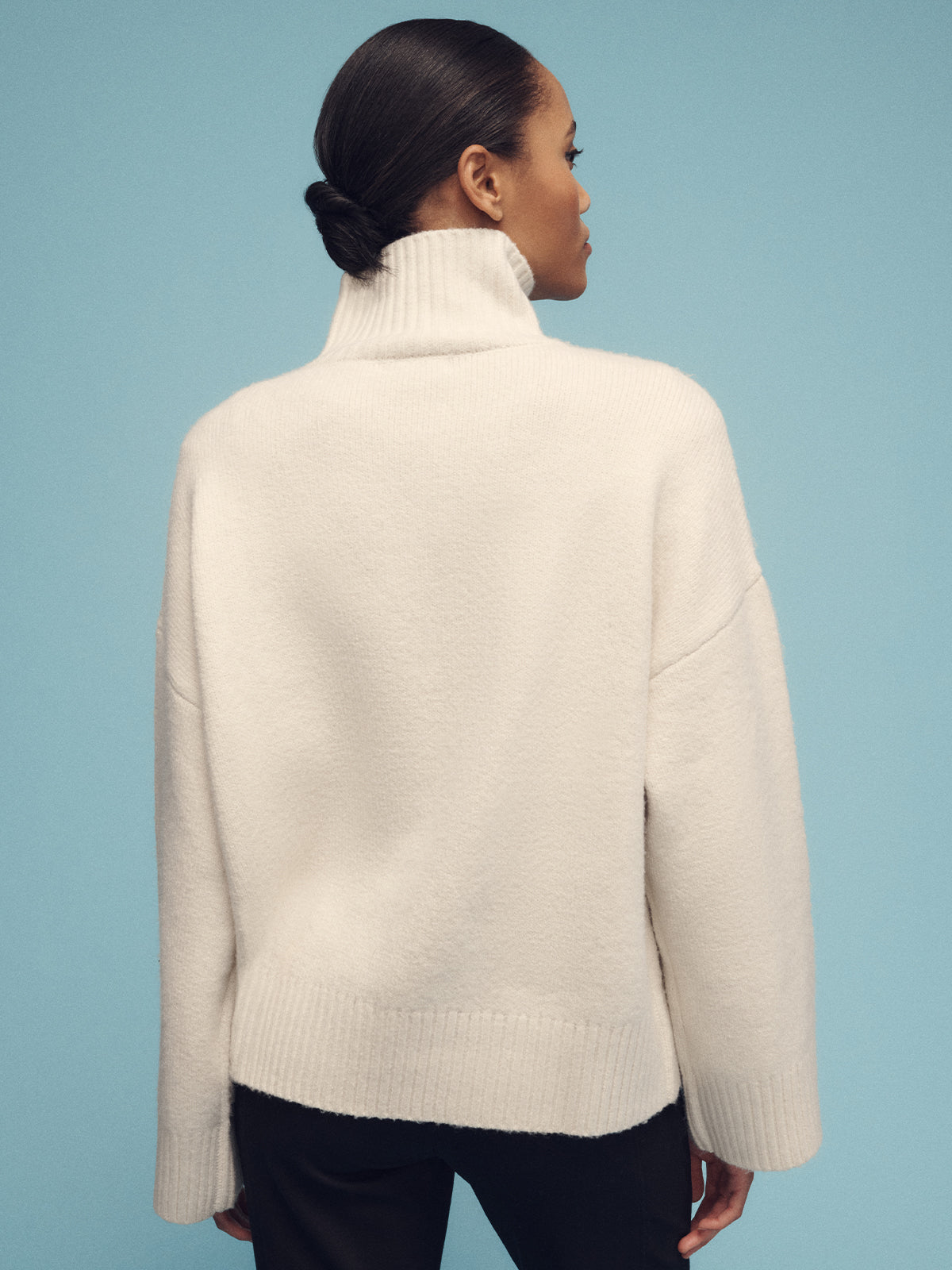 The individual stands against a pale blue background, facing away from the camera. They wear Sanctuary Clothing's 1/4 zip cashmere-like sweater in chalk with long sleeves and black pants. Their hair is pulled back into a neat bun.