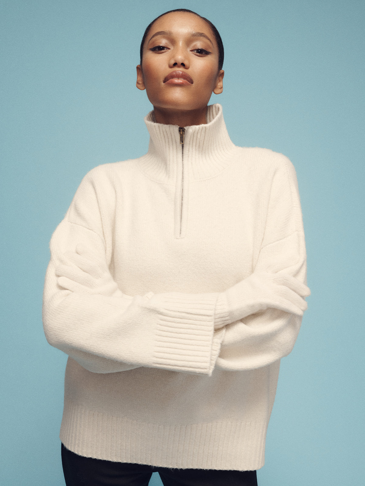 A person poses confidently against a blue backdrop, showcasing the chic, casual style of Sanctuary Clothing's 1/4 Zip Cashmere-Like Sweater in chalk, paired with black pants. The sweater features a cream-colored high-neck design with a half-zip and ribbed cuffs.