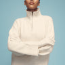 A person poses confidently against a blue backdrop, showcasing the chic, casual style of Sanctuary Clothing's 1/4 Zip Cashmere-Like Sweater in chalk, paired with black pants. The sweater features a cream-colored high-neck design with a half-zip and ribbed cuffs.