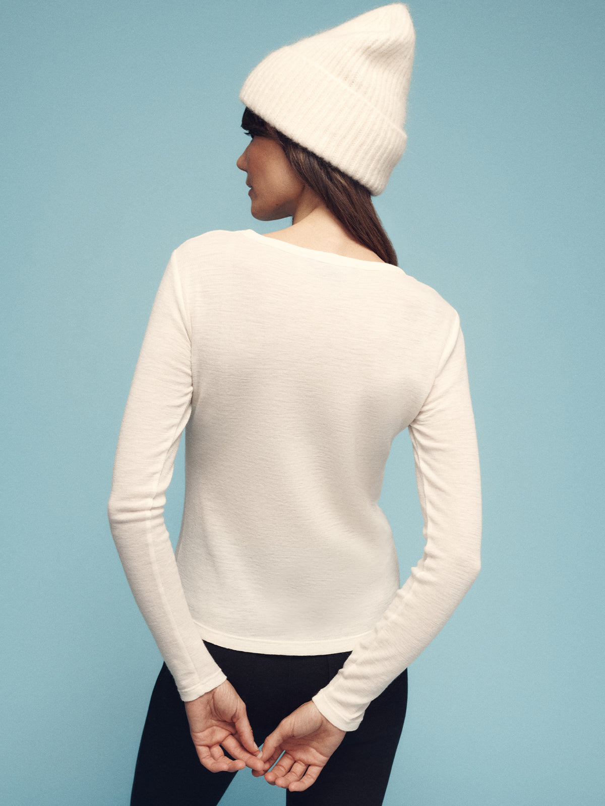 A person wearing a Sanctuary Clothing long sleeve wool perfect tee in chalk, black pants, and a white knit beanie is standing with their back to the camera against a light blue background. Their hands are clasped behind their back.