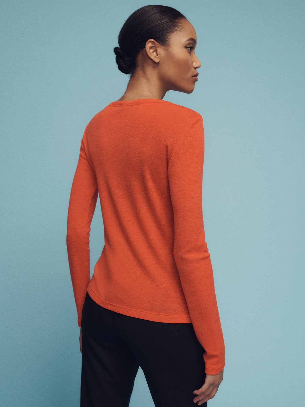 With their hair tied back, the person is facing away while wearing Sanctuary Clothing's "long sleeve wool perfect tee ski orange" and black pants against a plain blue background.