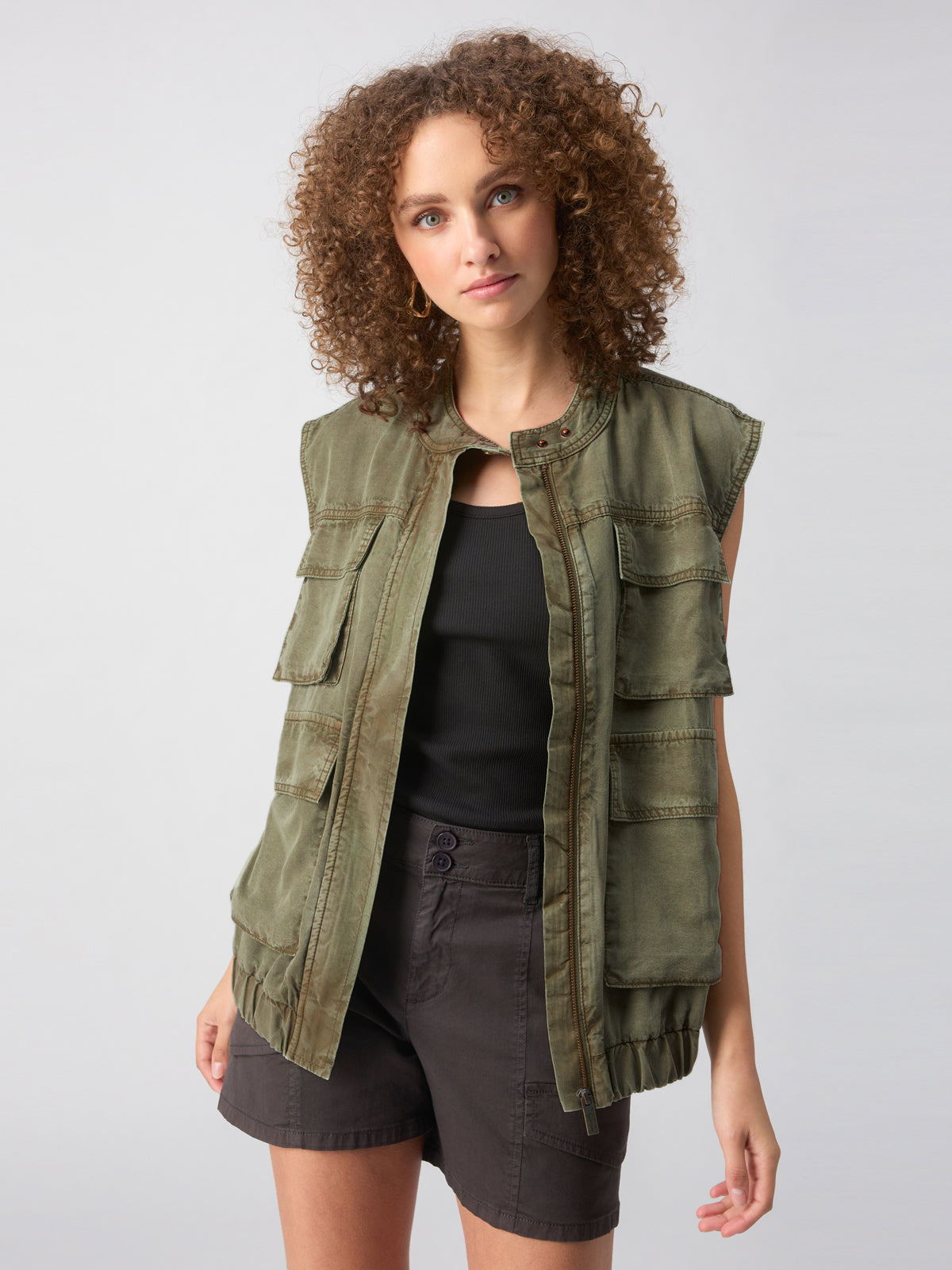 A person with curly hair, wearing the Field Utility Vest in Washed Olive by Sanctuary Clothing over a black tank top, is posing with their left hand in the pocket of dark shorts. The background is plain and light-colored.