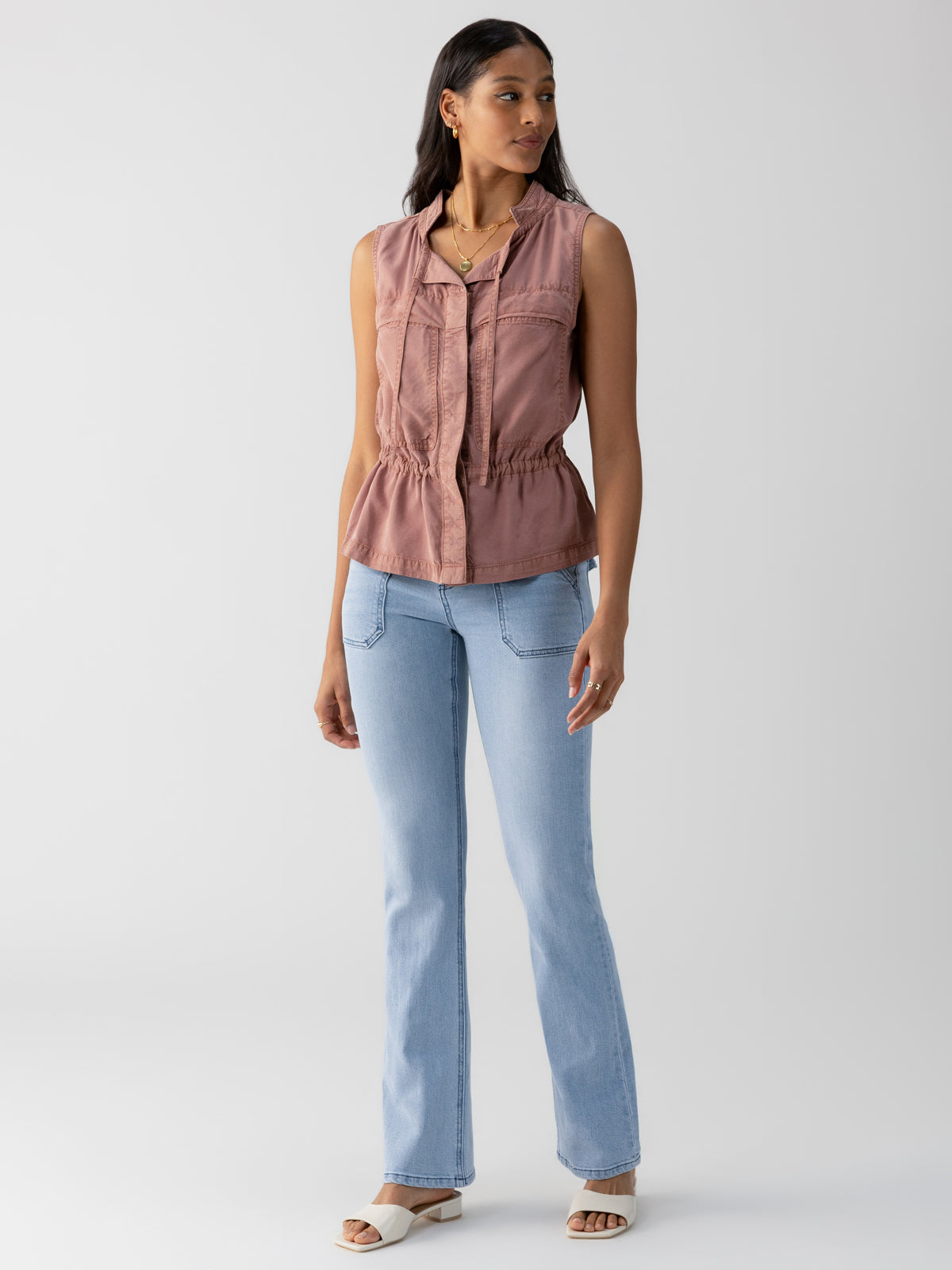 A person is standing against a plain white background, wearing a sleeveless Leah Surplus Vest in Washed Clay by Sanctuary Clothing, paired with light blue jeans and white sandals. They have long dark hair and are looking to their left.