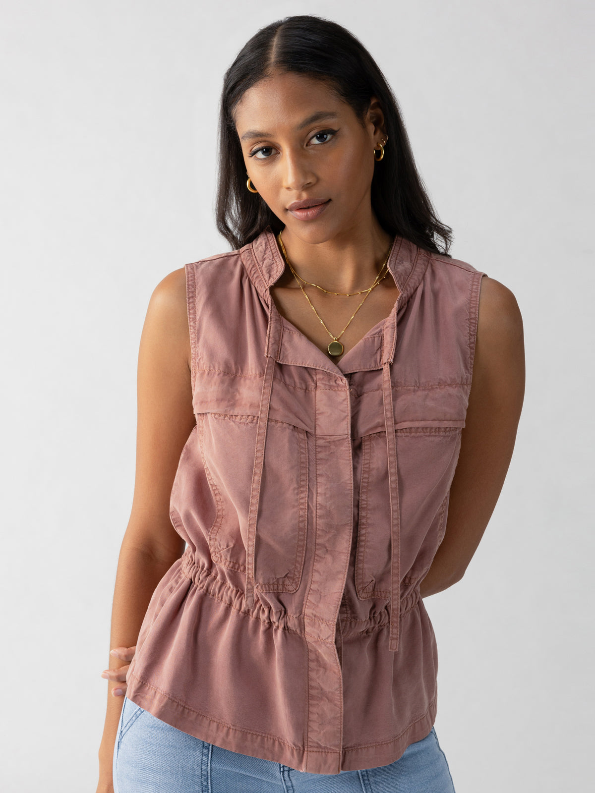 A woman with long dark hair is wearing the Leah Surplus Vest in Washed Clay by Sanctuary Clothing, paired with blue jeans. She has a serene expression and is accessorized with hoop earrings and layered necklaces. The background is plain and light-colored.