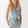 A person with long, curly hair is wearing the sleeveless Lora Surplus Vest in Eucalyptus by Sanctuary Clothing, featuring button details and a drawstring waist. They have their hands in their pockets and are posing confidently against a plain backdrop.