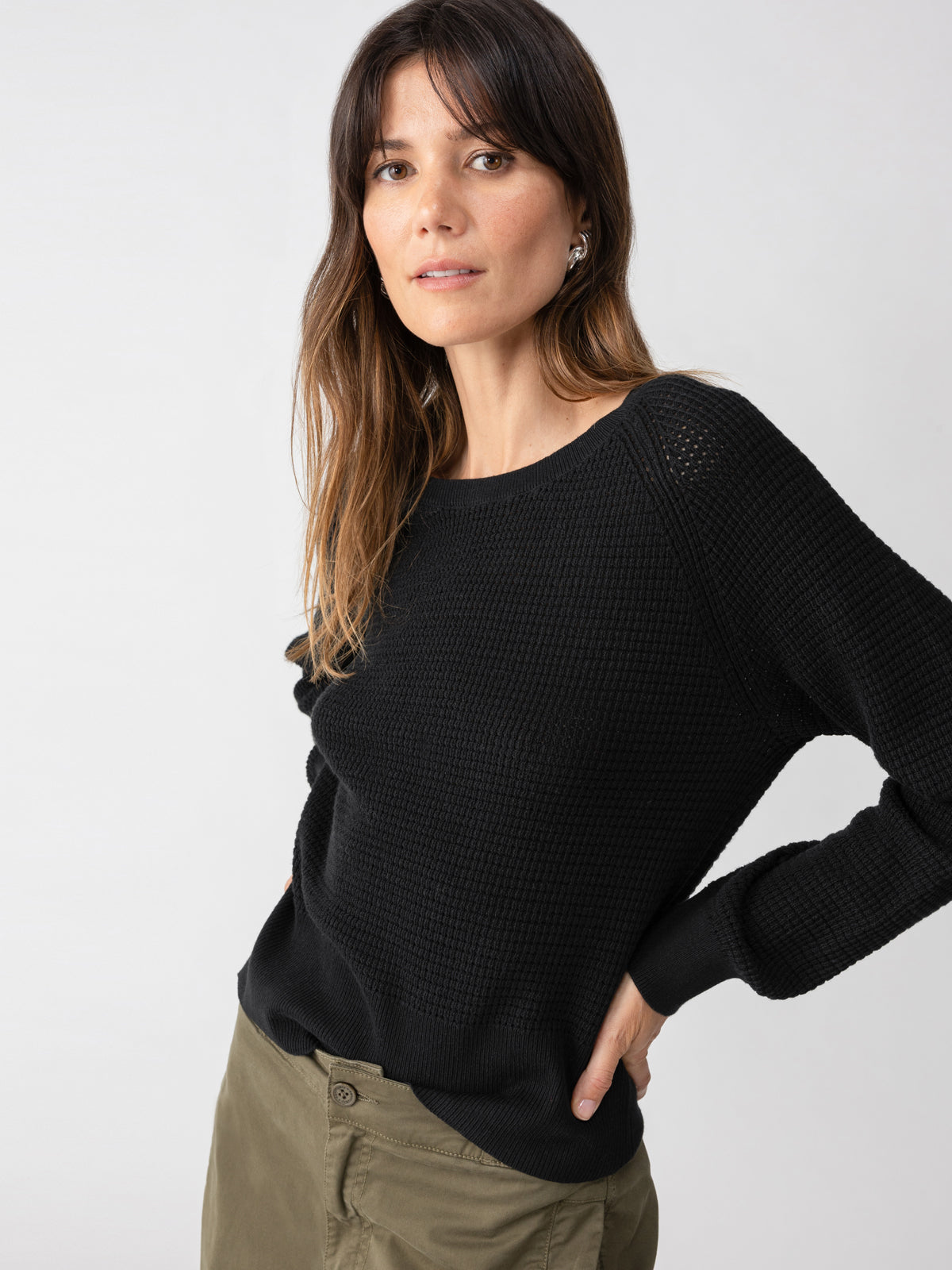 A woman with long brunette hair is wearing the Everyday Openwork Sweater in black by Sanctuary Clothing and khaki pants. She is standing against a neutral background, looking towards the camera with a relaxed expression, with one hand in her pocket.