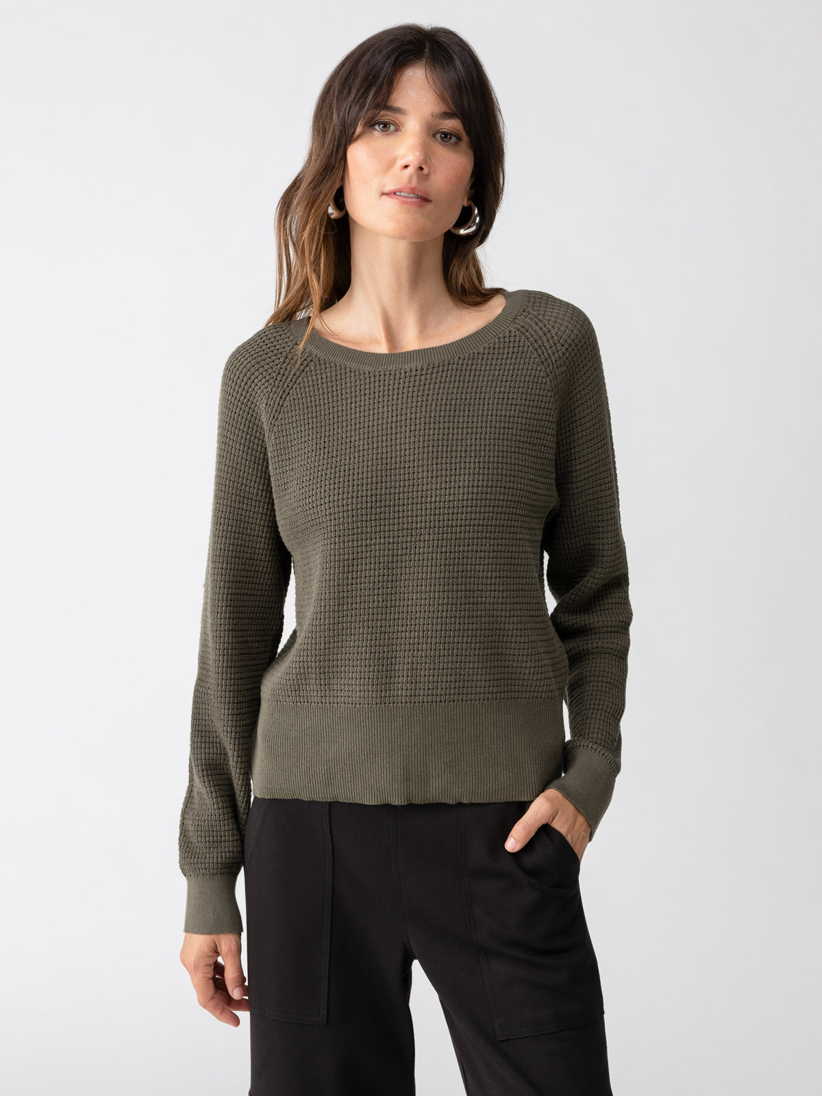 A person with long, brown hair is wearing the Everyday Openwork Sweater Kalamata by Sanctuary Clothing paired with black pants. With one hand in their pocket, they look confidently at the camera against a plain white background.