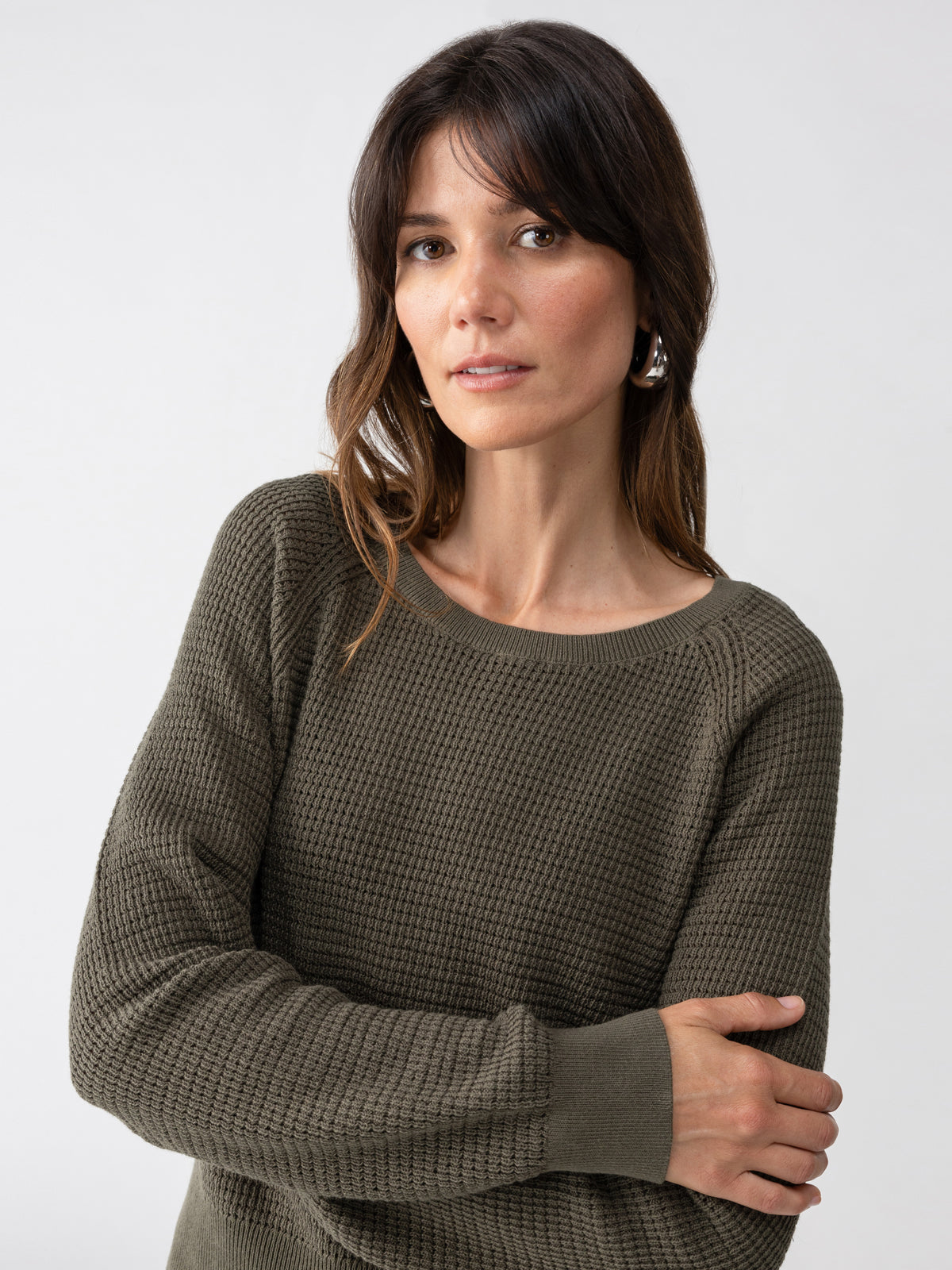 A woman with long dark hair and bangs, wearing the Everyday Openwork Sweater Kalamata by Sanctuary Clothing, looks slightly to the side with a neutral expression on her face. She has one arm crossed over her chest against a plain white background.