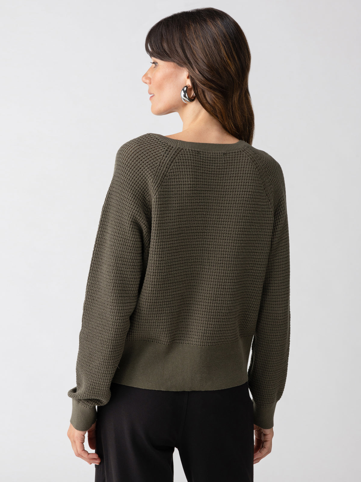 A person with long, dark hair stands facing away, showcasing the "Everyday Openwork Sweater Kalamata" by Sanctuary Clothing. The dark green, textured long-sleeve top features a ribbed hem and cuffs. They are also wearing black pants against a plain light grey background.