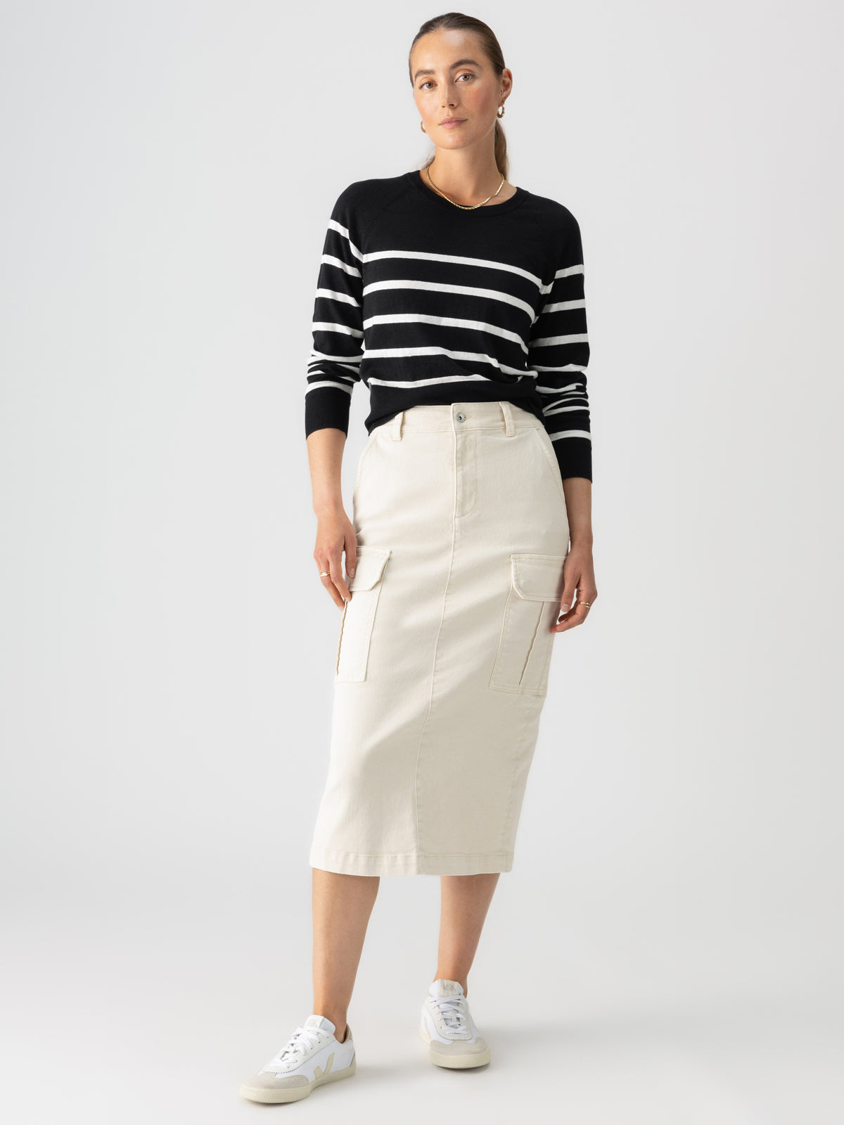 A person with long hair stands against a plain background, wearing the "All Day Long Sweater Black Chalk Stripe" from Sanctuary Clothing, paired with a beige, knee-length cargo skirt, and white sneakers. They have a relaxed pose with one hand in their skirt pocket and a neutral facial expression.