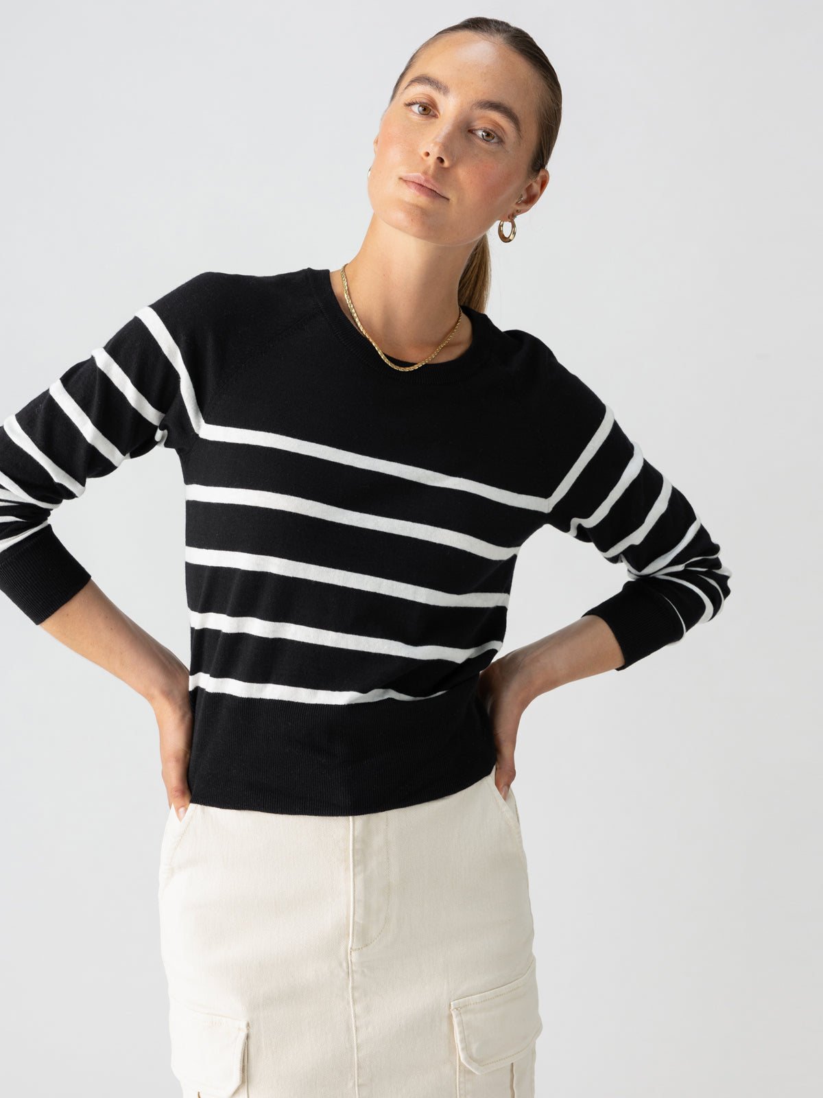 A woman stands confidently with her hands on her hips. She is wearing the "All Day Long Sweater Black Chalk Stripe" from Sanctuary Clothing, paired with a light-colored skirt featuring pockets. She accessorizes with gold hoop earrings and a gold necklace, and her hair is pulled back neatly. The background is plain white.