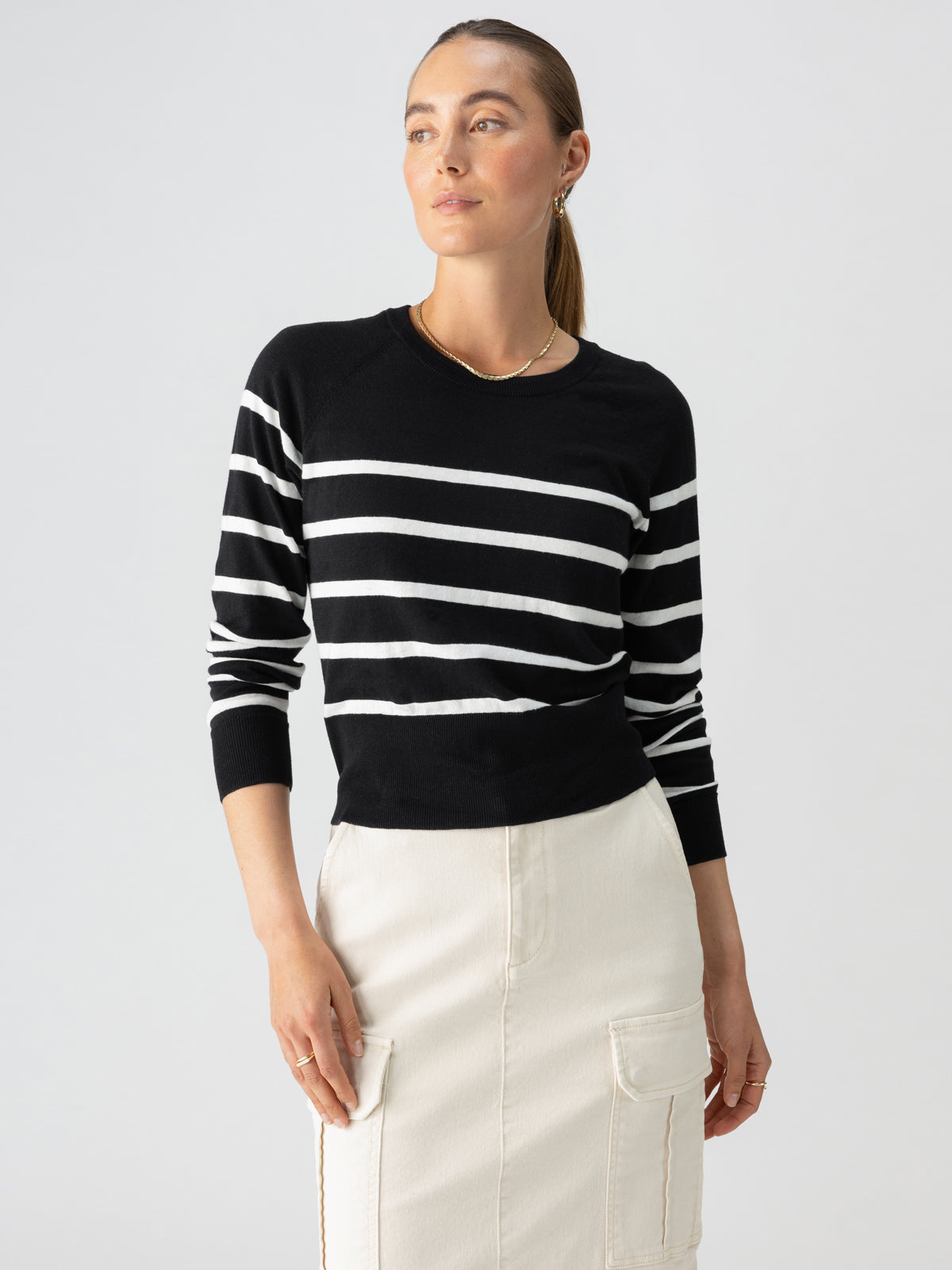 A person with long hair is gazing to the side while wearing the Sanctuary Clothing All Day Long Sweater in Black Chalk Stripe and a light-colored skirt with pockets. The individual, adorned with earrings and a necklace, stands against a plain white background.