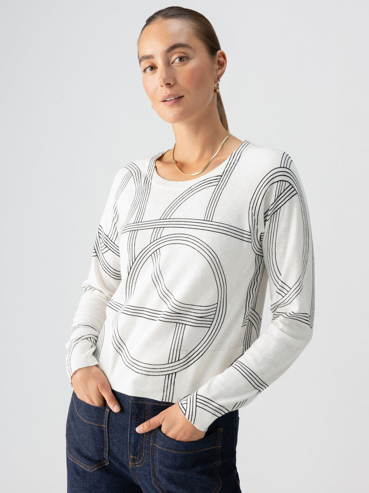 An individual with long hair tied back is wearing the "All Day Long Sweater Graphic Lines" by Sanctuary Clothing, paired with blue jeans. They are standing against a plain grey background, looking slightly off to the side with hands partially in their pockets.